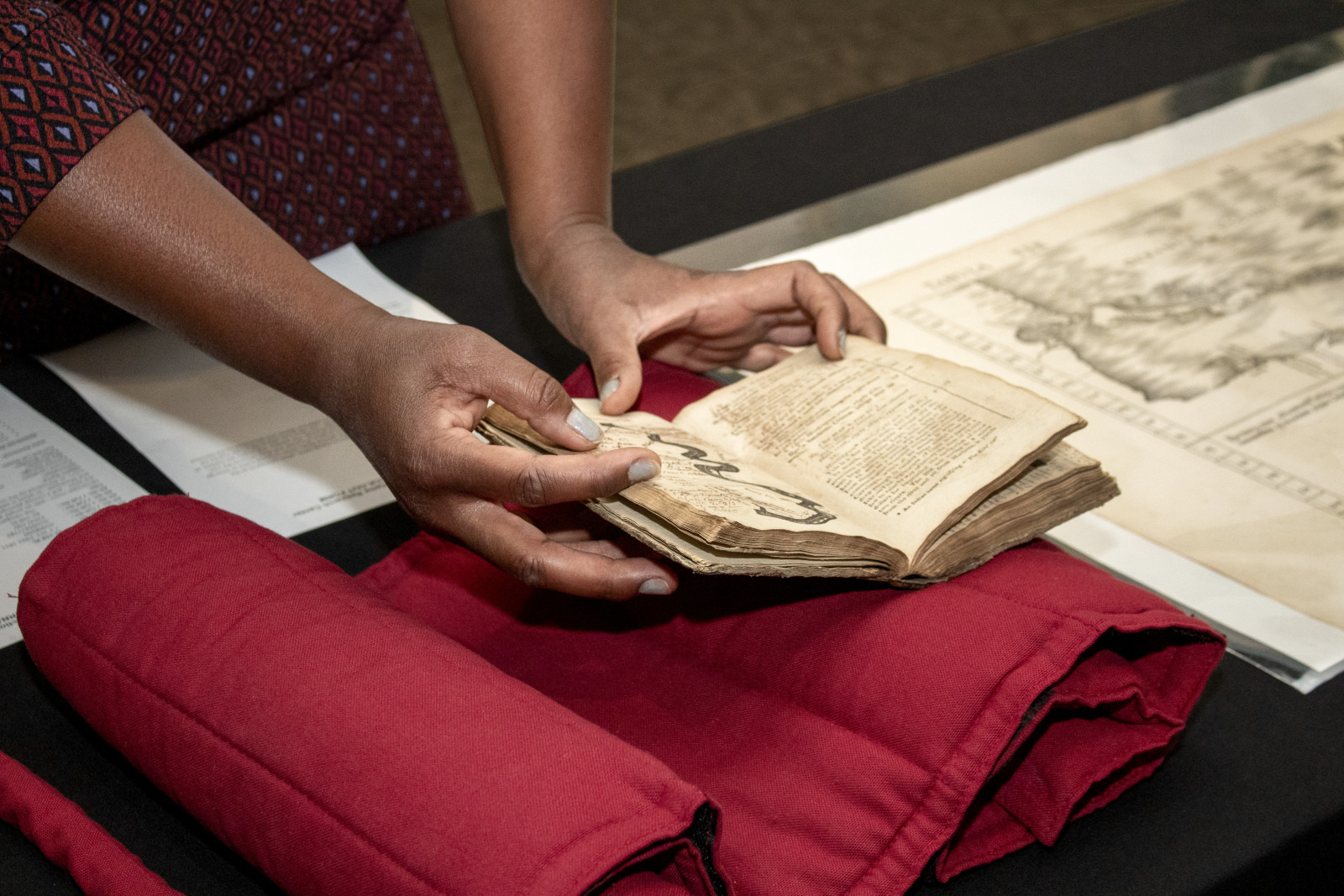 A person carefully handles an old, open book above a red fabric on a table. Nearby is a large, detailed map or illustration. The scene suggests an environment focused on historical documents or archival work.
