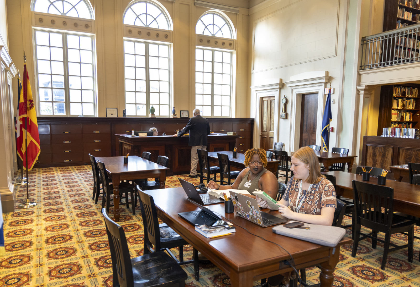 Two young women researchers discuss a book while sitting at a wooden table in the middle of a large library space.
