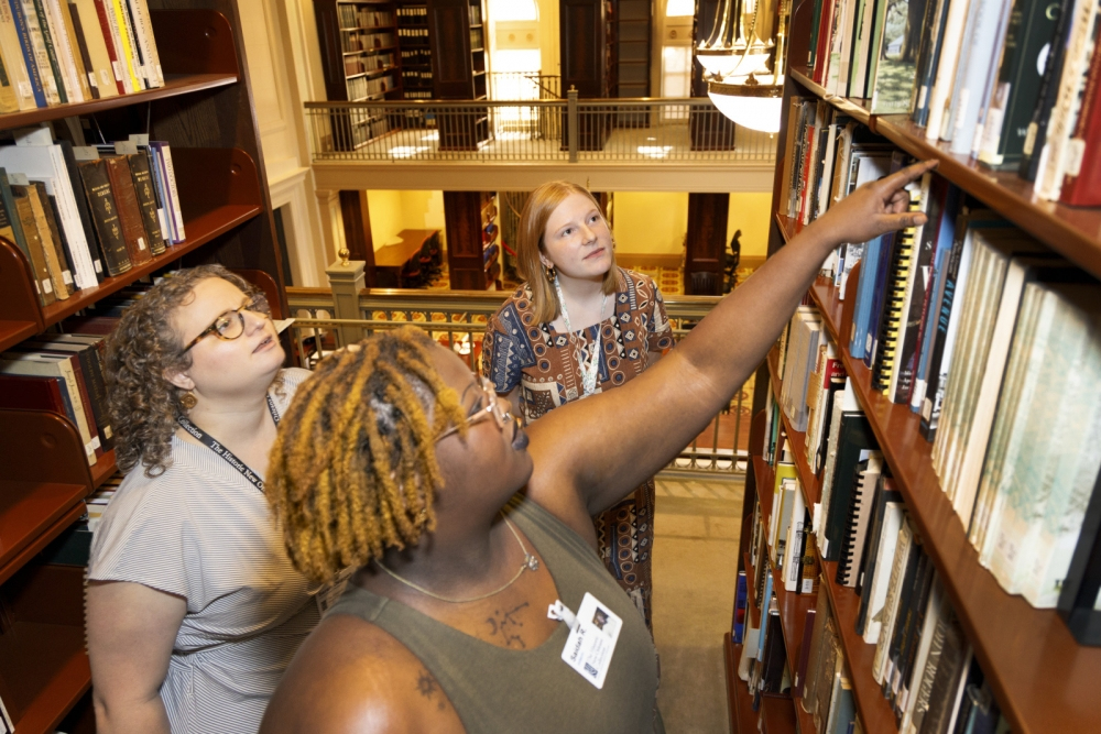 Three women are looking at books on a library shelf. One is reaching for a book, while the others watch. They stand in a well-lit library with rows of books and an ornate railing in the background.