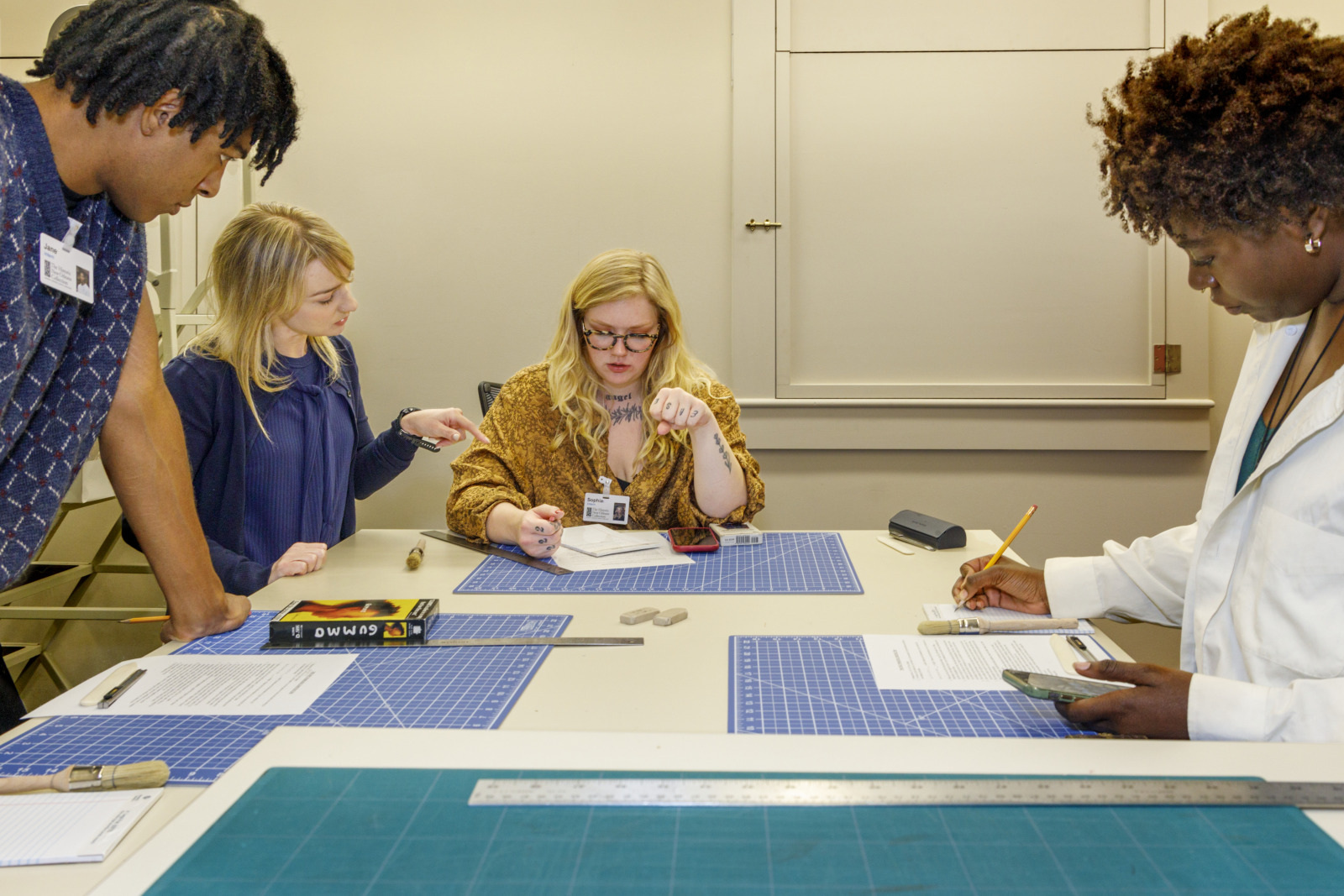Four people are gathered around a table with blue cutting mats, examining objects and taking notes. They appear to be collaborating on a project, possibly related to archaeology or art restoration, as they focus on small artifacts.