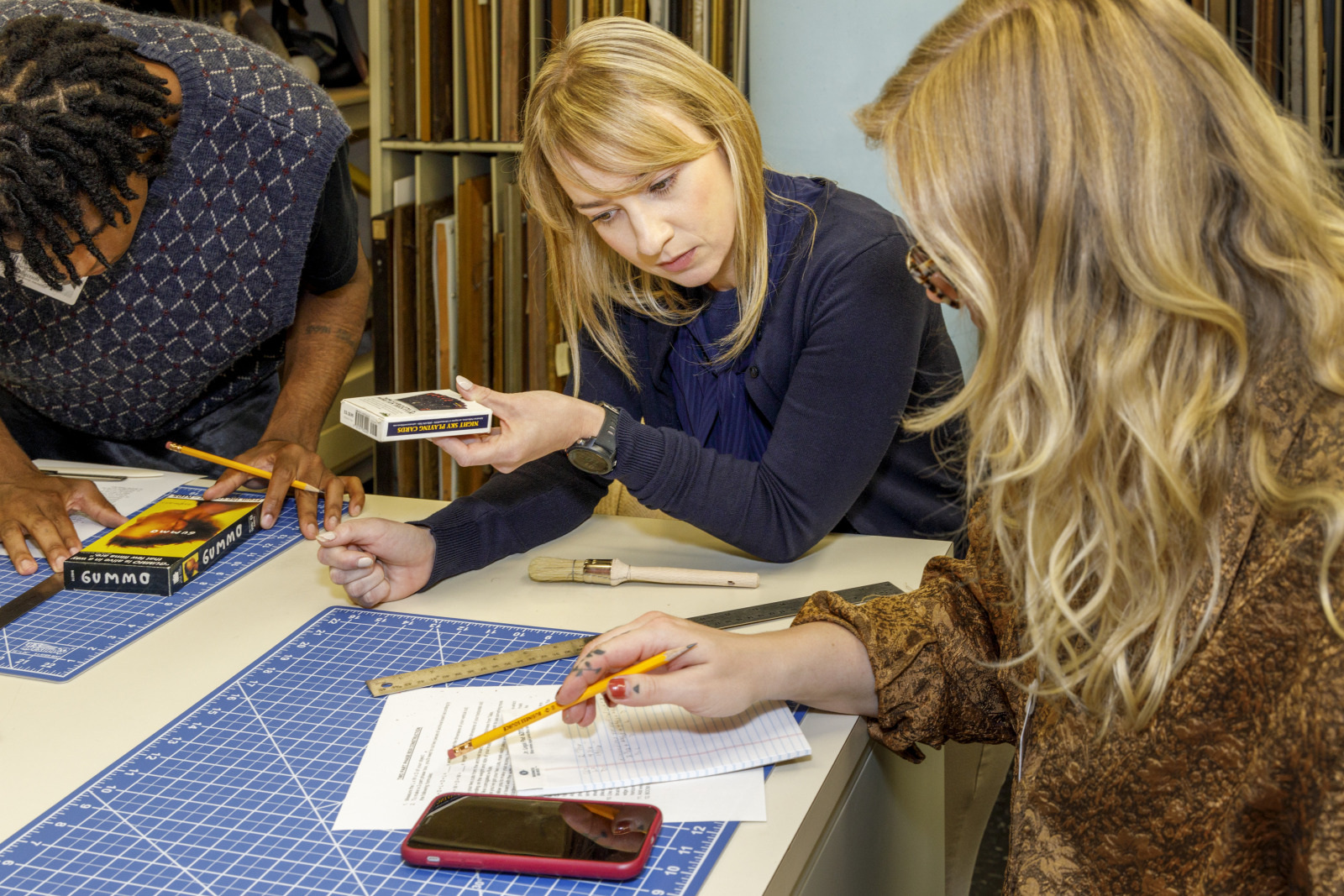 Three people work collaboratively at a table. One woman holds a color palette and papers, while the others engage with pencils. A blue cutting mat, a smartphone, and colored pencils are visible. Shelves with books are in the background.