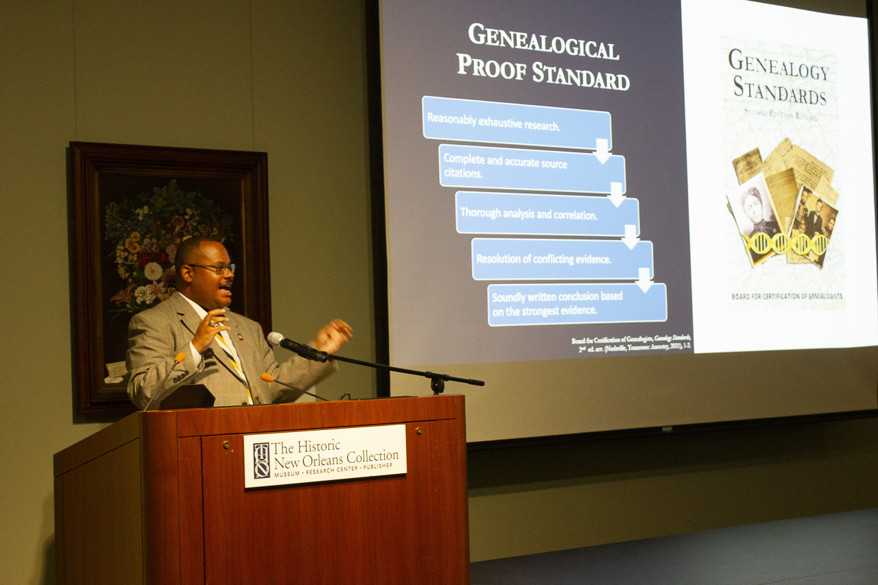 A man in a suit is giving a presentation at The Historic New Orleans Collection. The slide he presents outlines the Genealogical Proof Standard, featuring key steps and a book cover of Genealogy Standards.