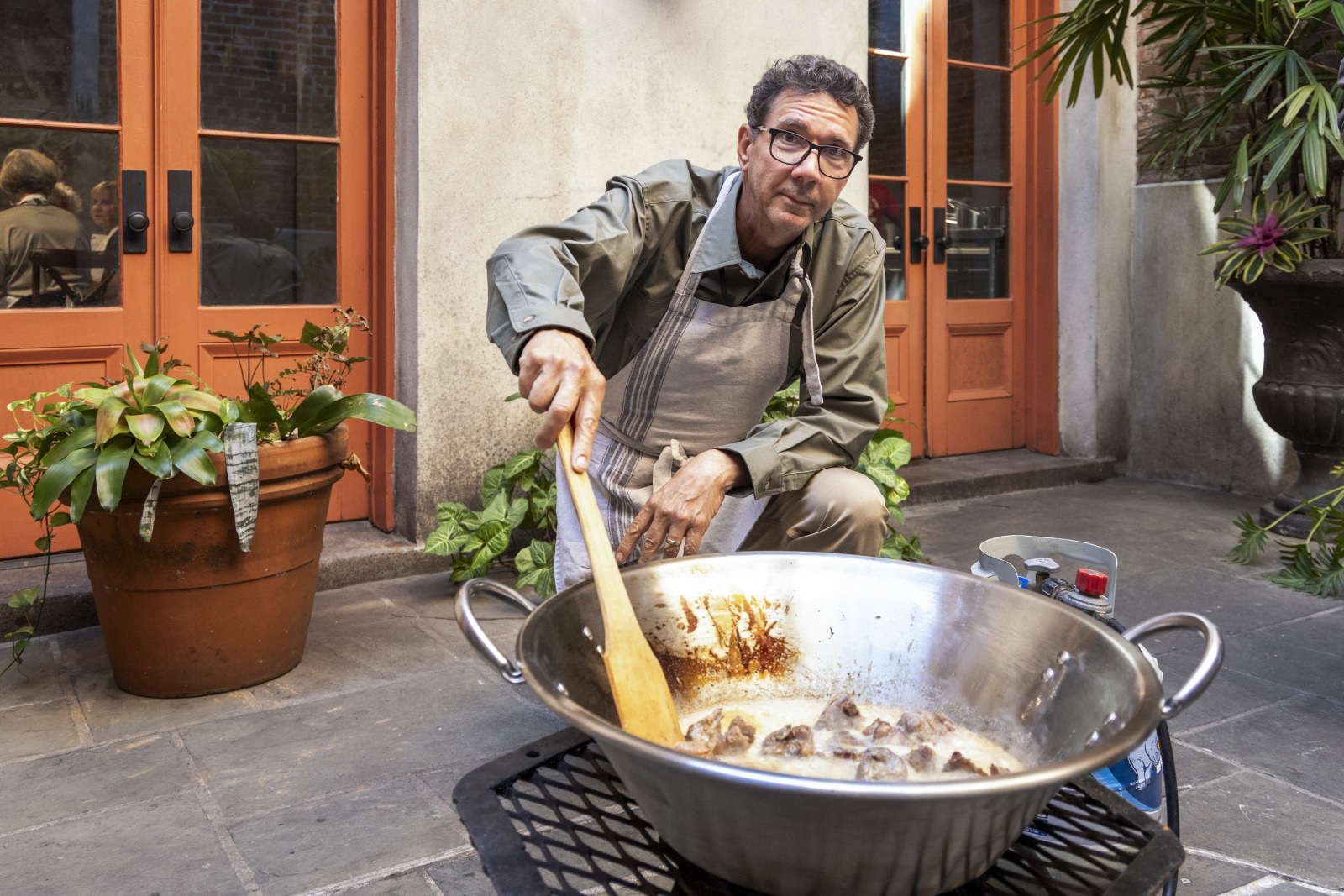 A man wearing glasses and an apron is cooking outdoors, stirring a large metal pan on a grill. The scene is set against a backdrop of orange-framed doors and potted plants on a patio.