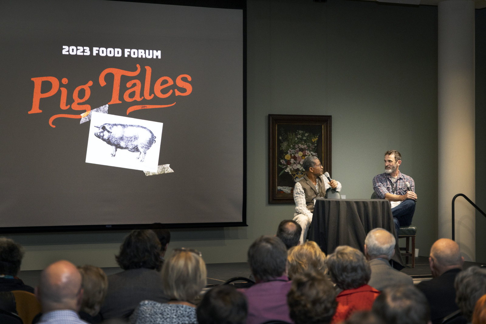 A speaker and a host sit on stage at the 2023 Food Forum event titled Pig Tales. The stage features a large screen with the event name and an illustration of a pig. An audience watches the presentation.