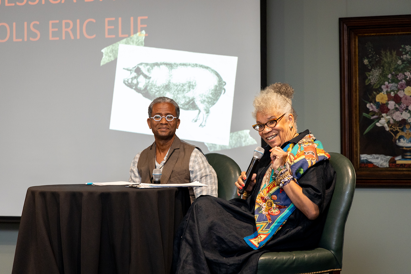 Two people are seated on stage for a discussion. One holds a microphone, smiling and wearing colorful glasses. A projection screen behind them displays text and an image of a pig. A painting is partially visible on the wall.