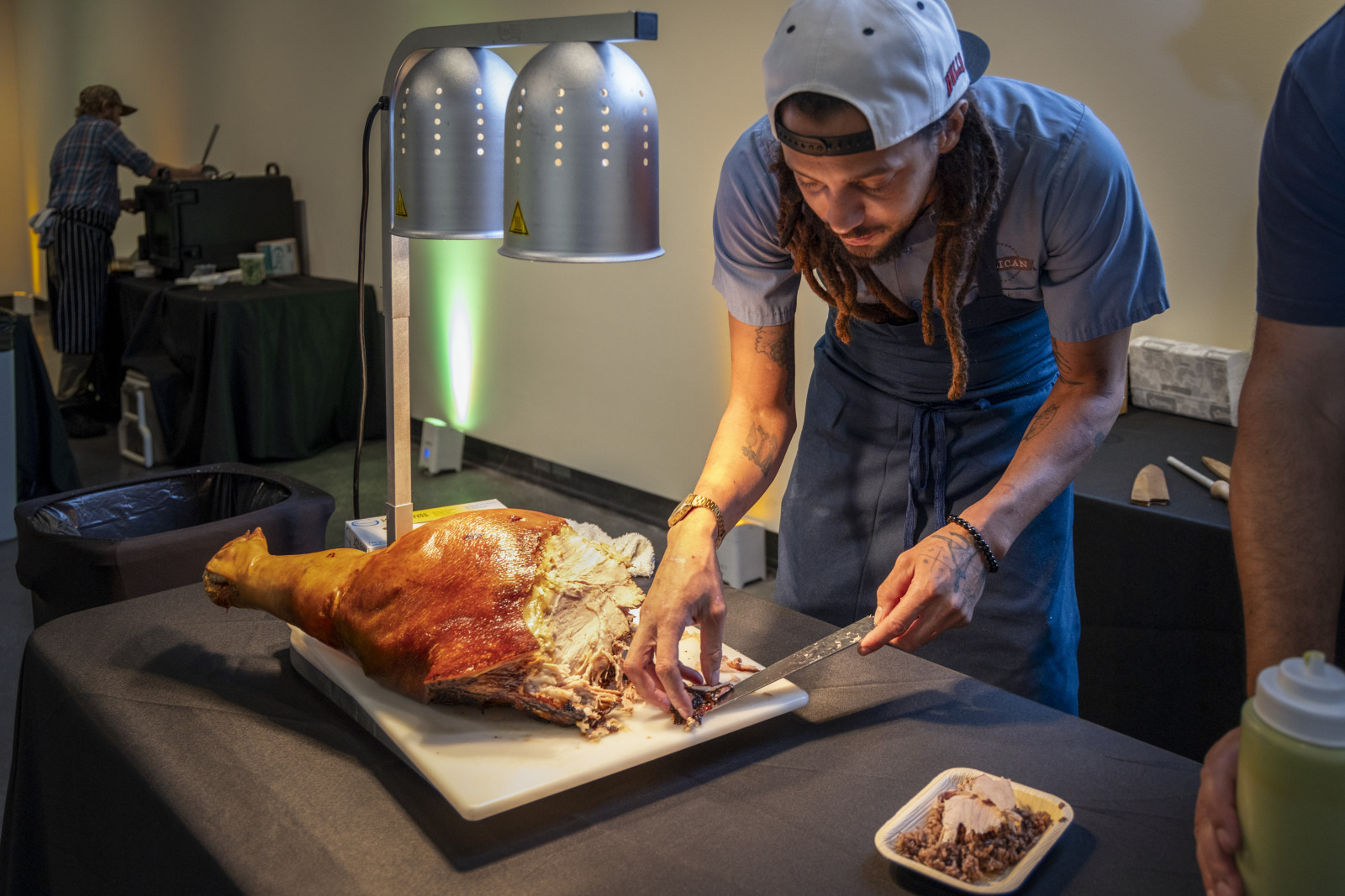 A chef with long dreadlocks and a backward cap carves a large piece of roasted meat under a heat lamp at a food station. A tray with slices of meat and a bottle of sauce are nearby. Another person works in the background.