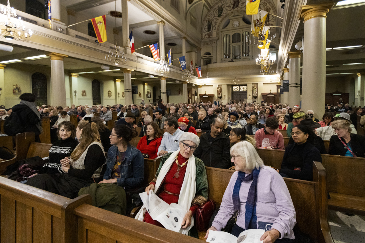 A large crowd of people seated in pews inside a grand church with flags hanging from the ceiling. The interior features ornate architecture and chandeliers. Some individuals are reading from booklets.