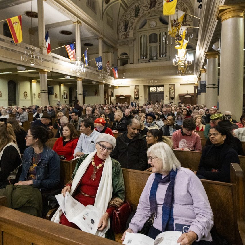 A large congregation sits in wooden pews inside a church, decorated with colorful flags. People of various ages are engaged, reading programs or conversing. The architecture features high ceilings and columns.