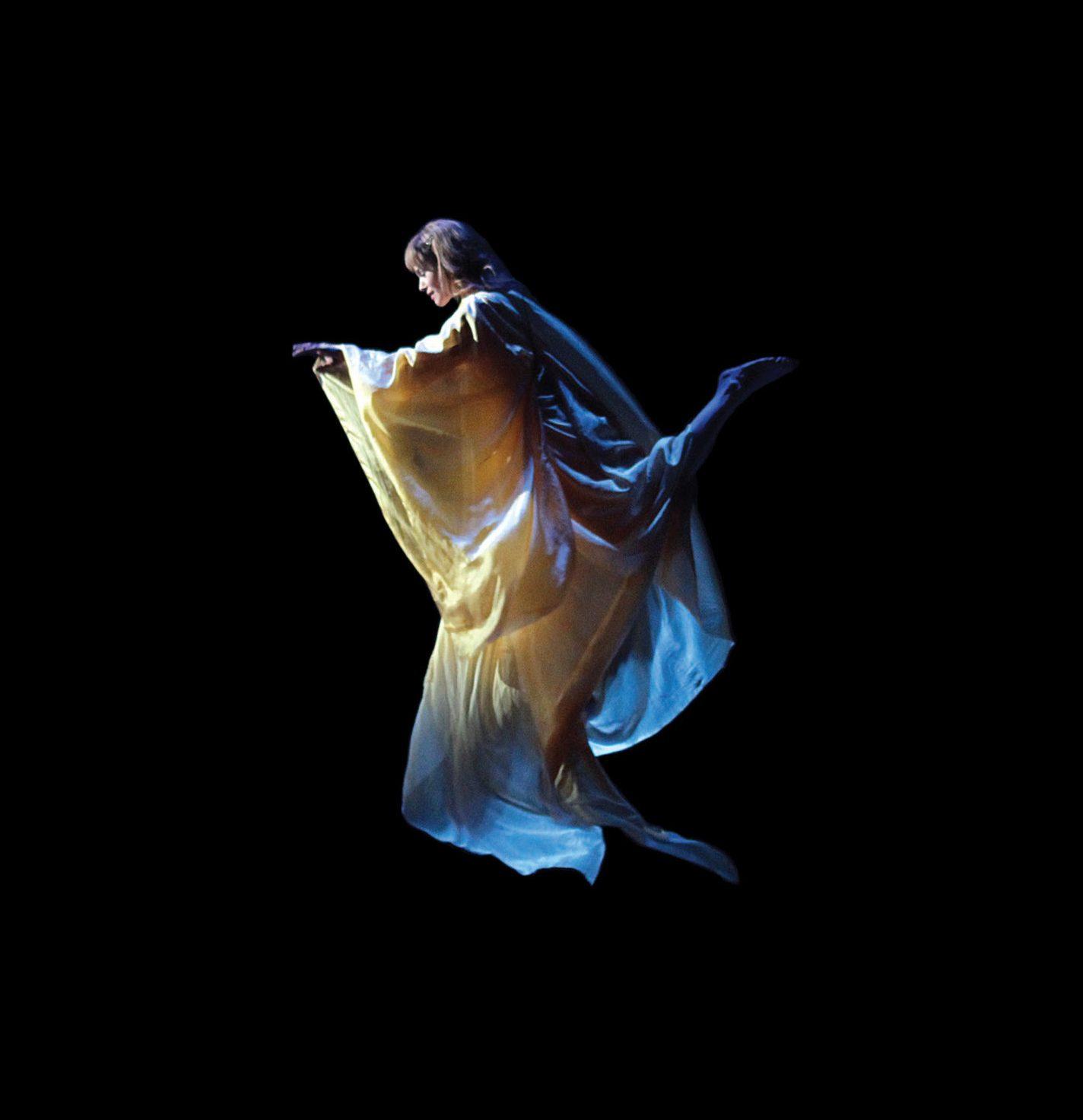 A dancer gracefully leaps mid-air against a dark background, wearing a flowing, golden robe that billows around them. The lighting highlights the fabric, creating an ethereal effect.