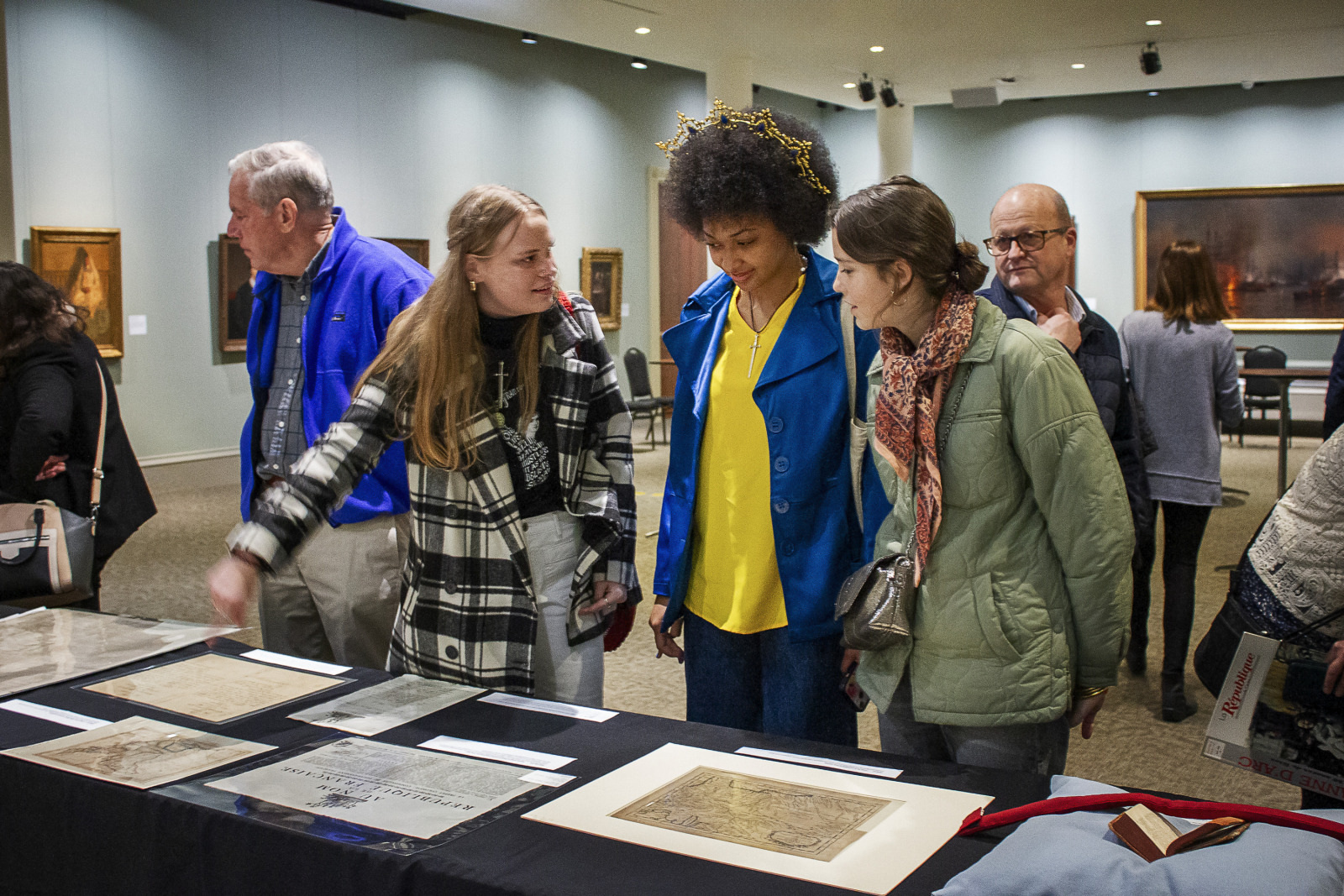 A group of people in a museum examines historical documents displayed on a table. Several artworks hang on the walls behind them. They appear engaged and interested in the items.