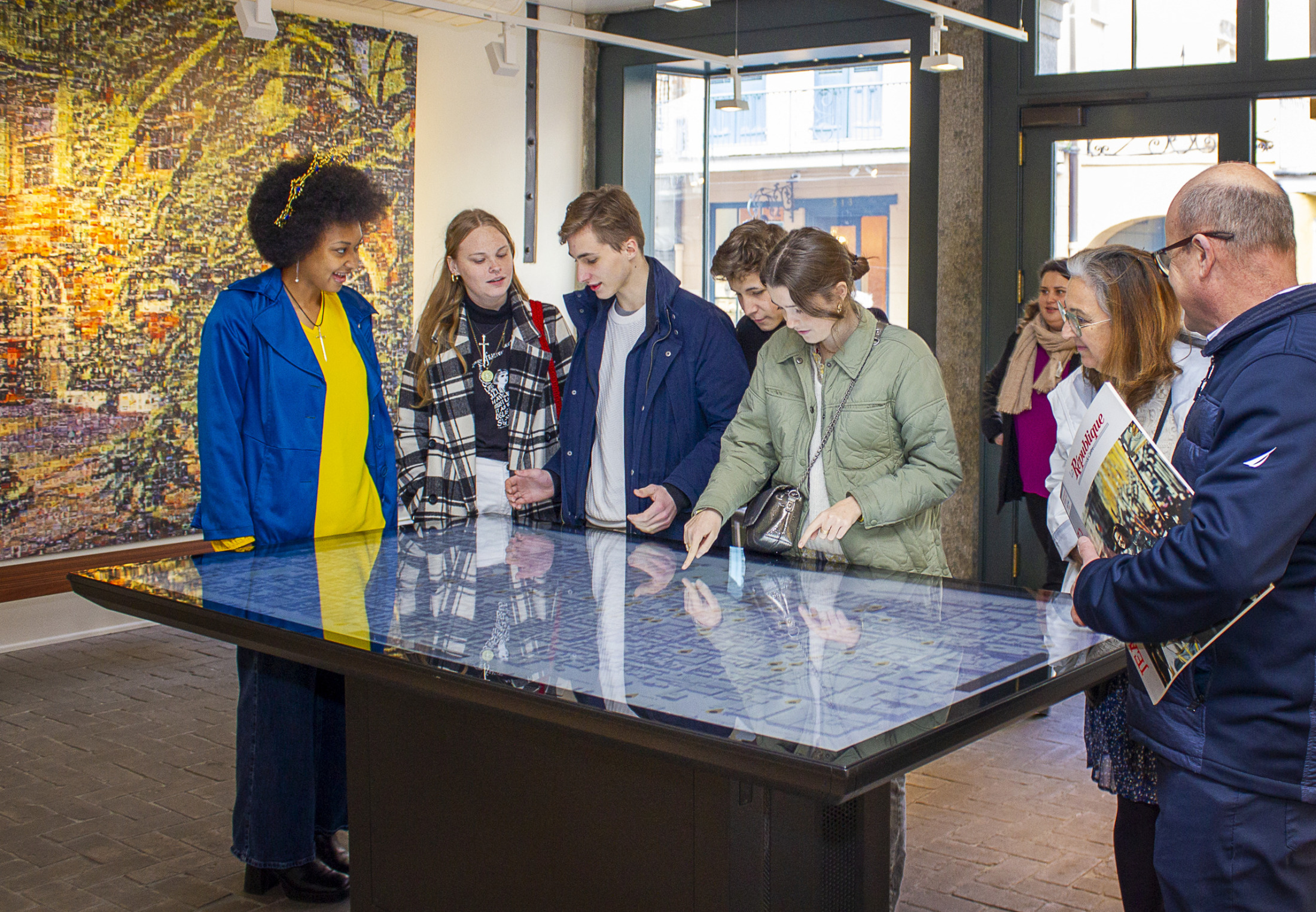 A group of people in a museum examines a map on a digital table. Several artworks hang on the walls behind them. They appear engaged.
