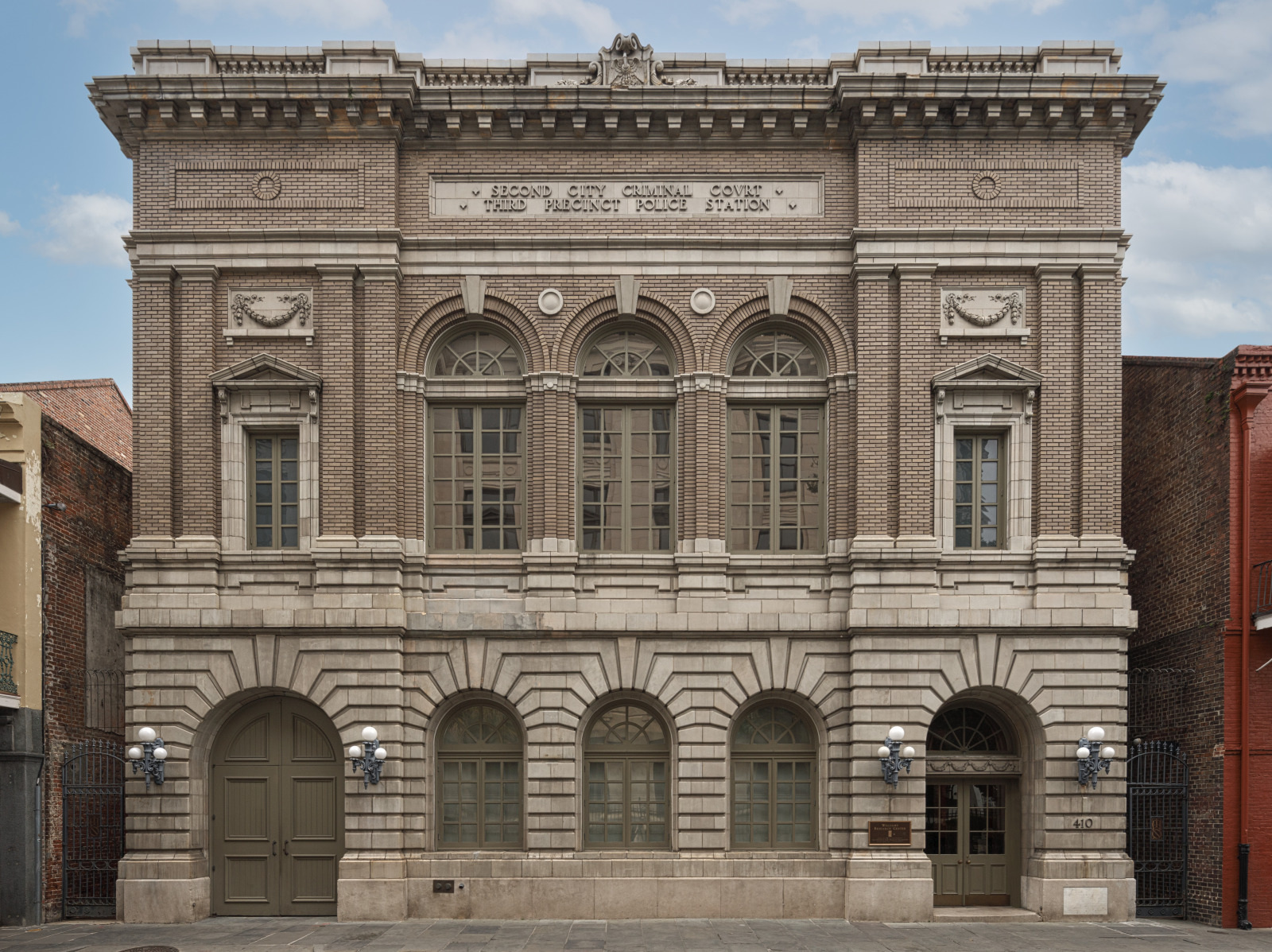 A historic brick building with tall, arched windows and ornate stone details. The facade features a central sign with engraved text. Large doors and lantern-style lights flank the entrance. The structure is framed by neighboring buildings.