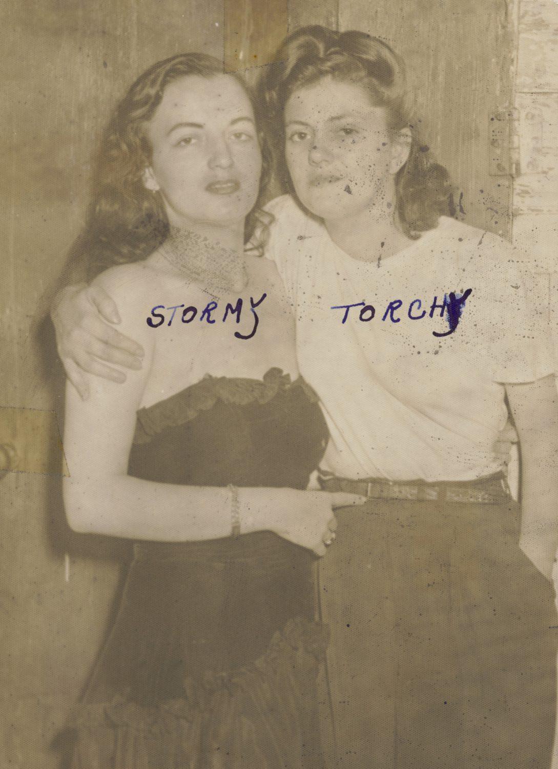 A vintage sepia-toned photo of two women. The left woman, labeled Stormy, wears a dark strapless dress. The right woman, labeled Torchy, wears a white blouse and dark pants. They pose together affectionately, with their arms around each other.