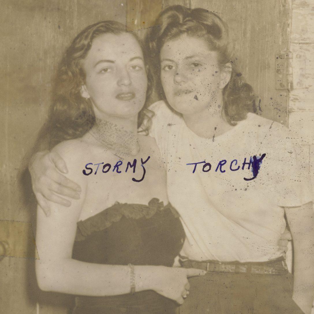 Two women pose closely together in a vintage black and white photo. One wears a strapless dress, and the other is in a white shirt. The names Stormy and Torchy are handwritten above them.