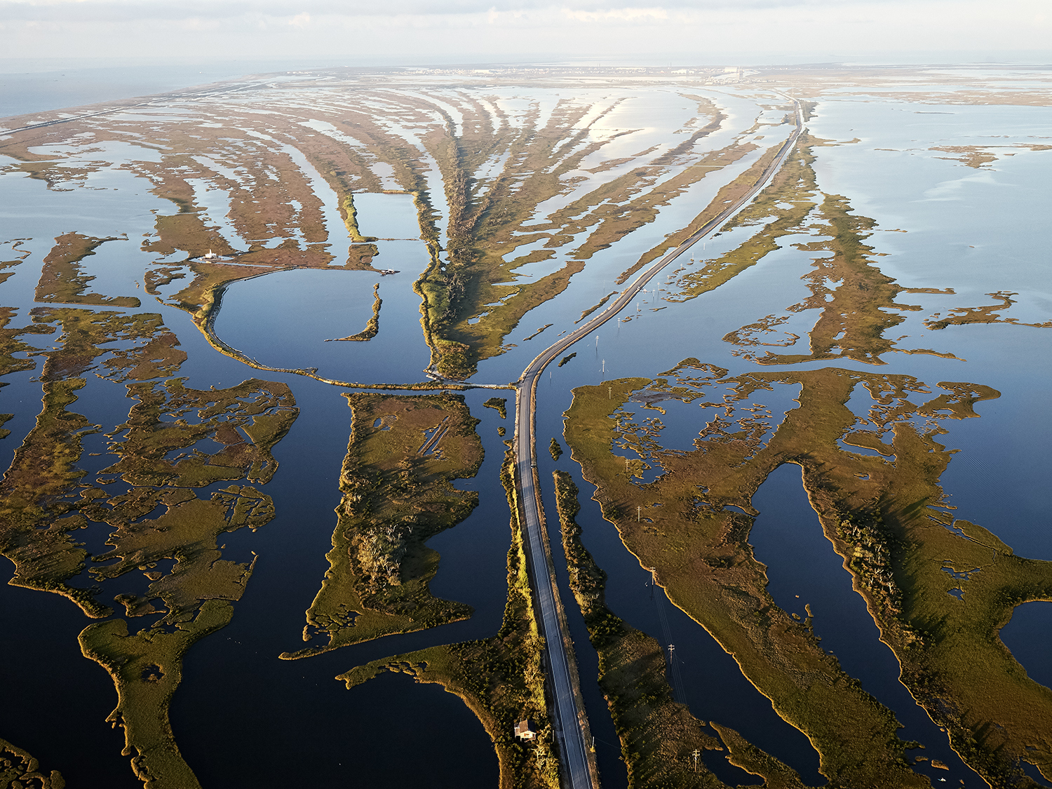 Aerial viewo f the eroding marsh and open water surrounding Louisiana Highway 1 in Jefferson Parish, showing a winding road cutting through a marshy landscape with numerous water channels and patches of green vegetation. The sky is clear, reflecting on the waters surface.