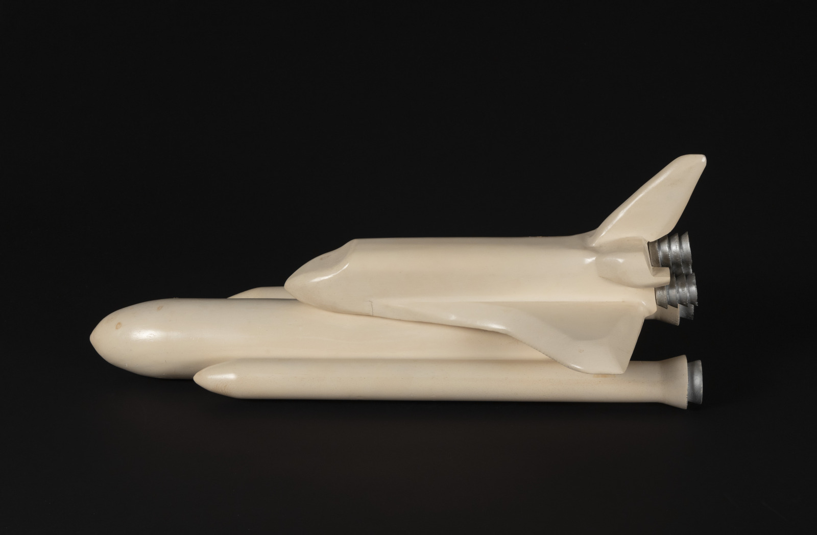 A side profile view of a faded white model of a space shuttle, connected to its large fuel tank and rocket boosters. The model is "blank" without details on any features except for the rocket nozzles and is set against a black background.