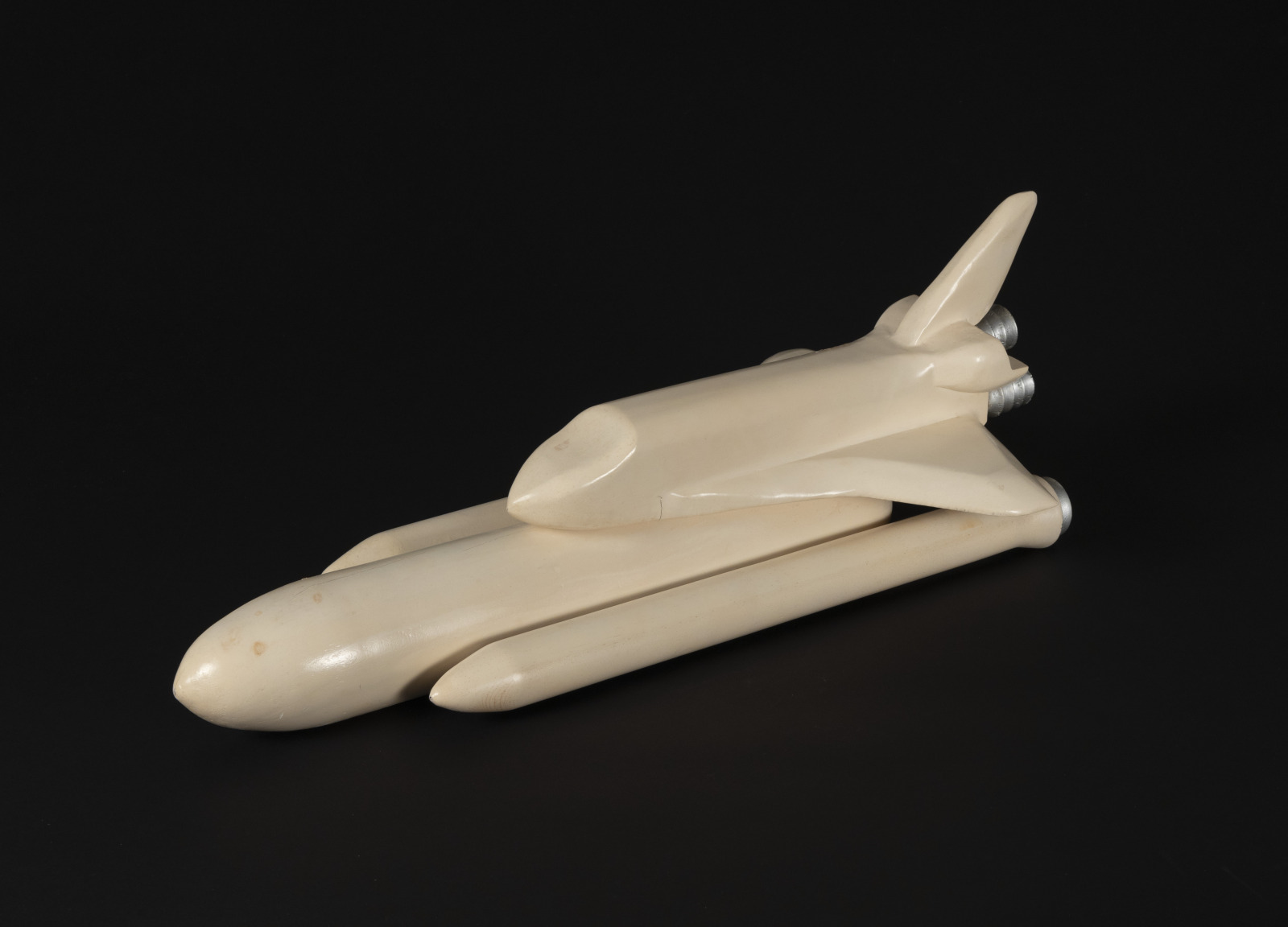 A view from above of a faded white model of a space shuttle, connected to its large fuel tank and rocket boosters . The model is "blank" without details on any features except for the rocket nozzles and is set against a black background.