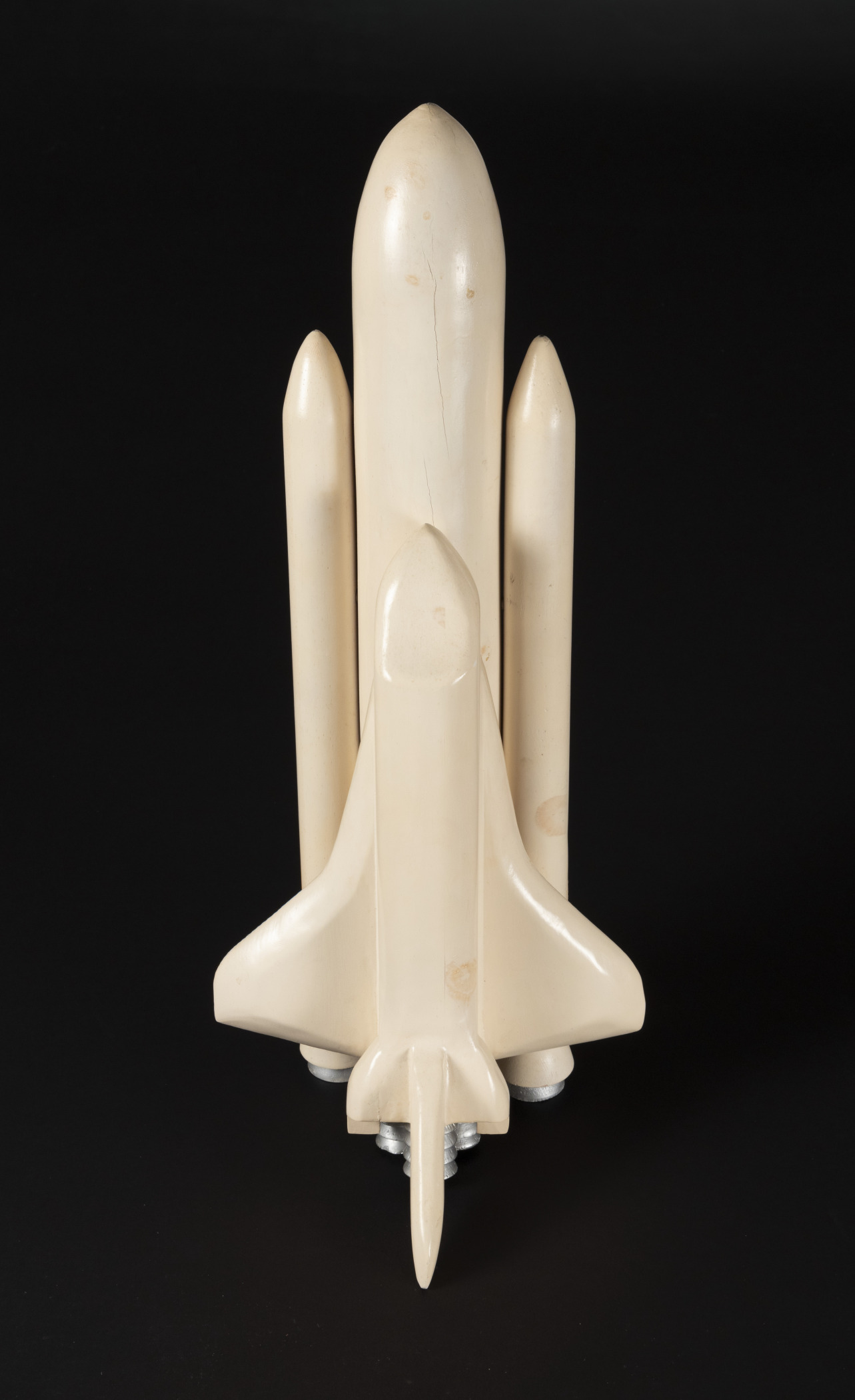 A view from above of a faded white model of a space shuttle, connected to its large fuel tank and rocket boosters. The model is "blank" without details on any features except for the rocket nozzles and is set against a black background.