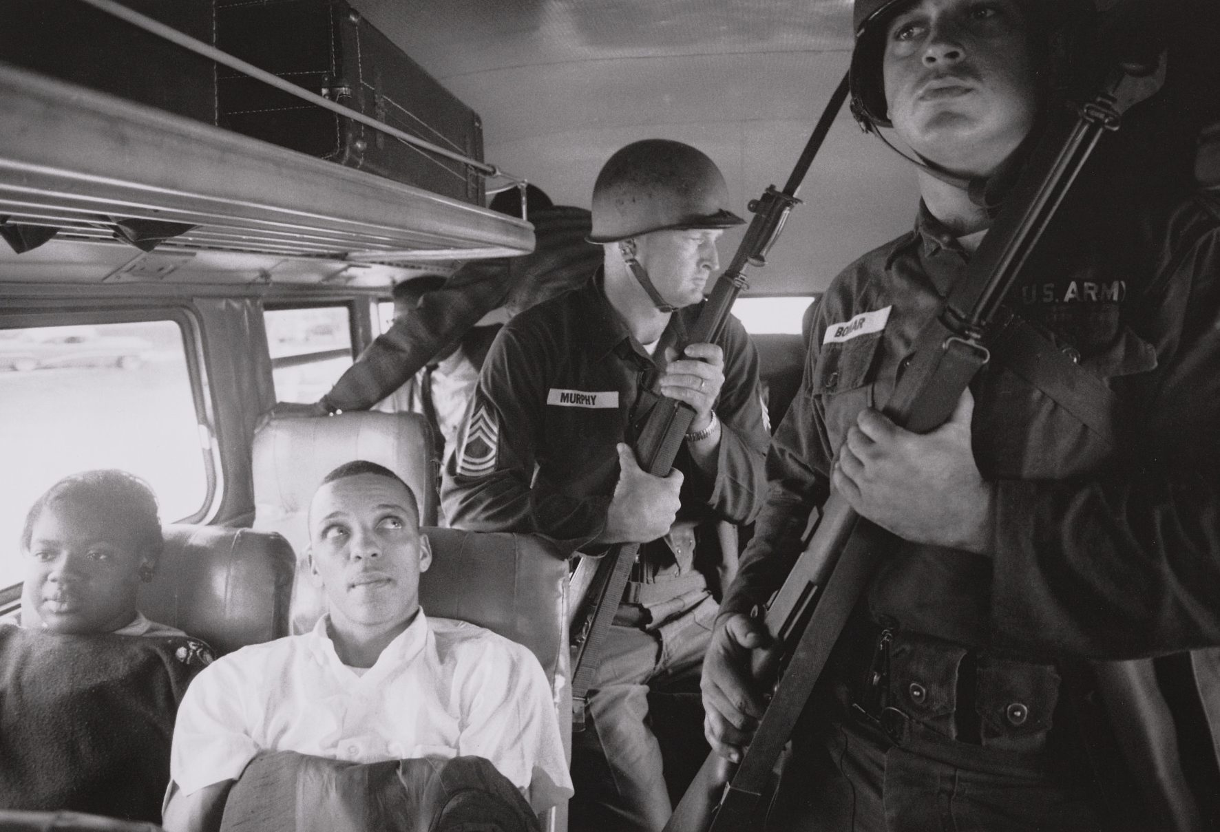 A black and white photo shows Freedom Riders Julia Aaron Humbles and David Dennis sitting near two armed National Guardsmen on a bus from Montgomery, Alabama to Jackson, Mississippi in May of 1961.