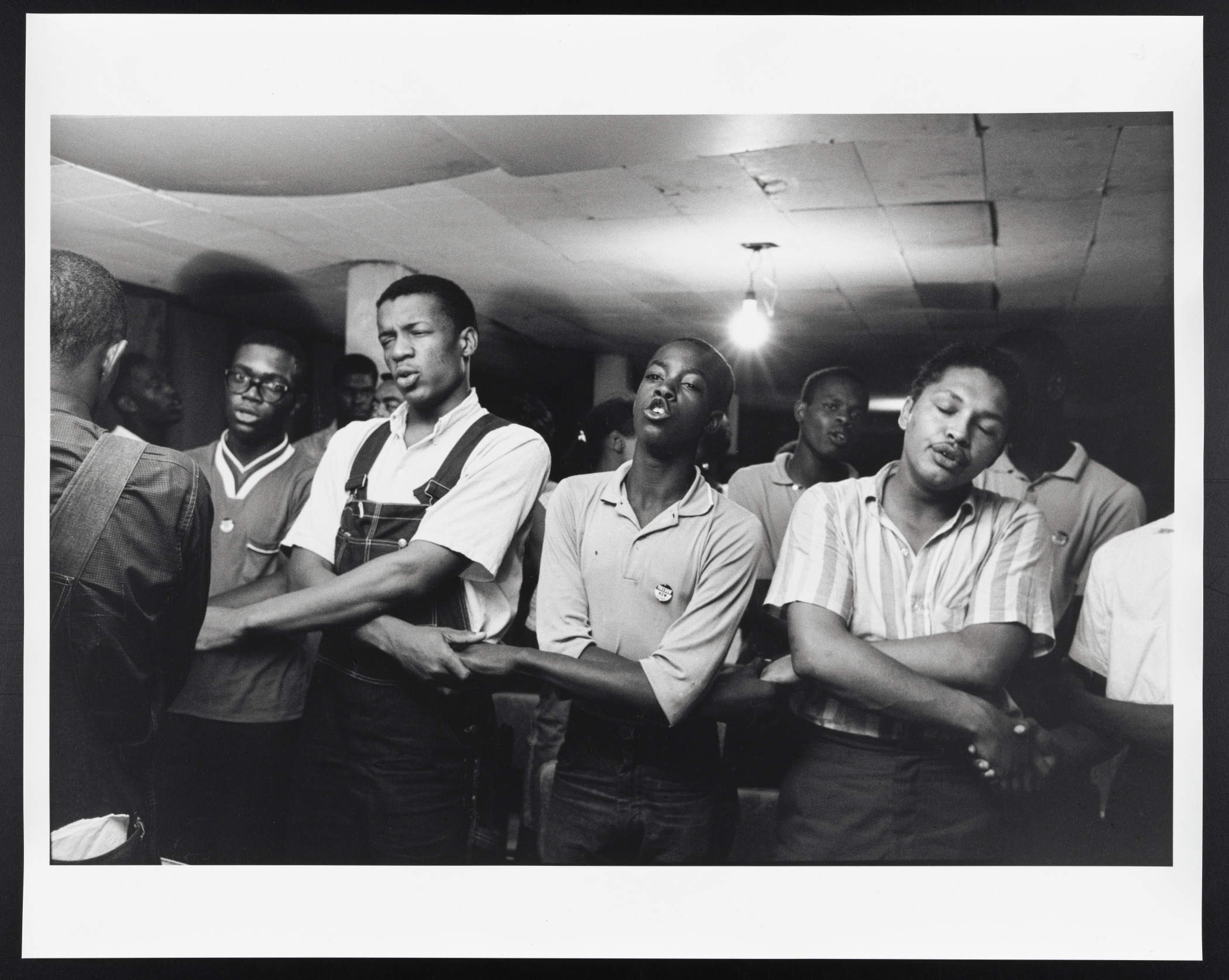 A black and white photograph shows Jerome Smith leading a group of New Orleans CORE members, their arms intertwined, singing “We Shall Overcome,” in 1963.