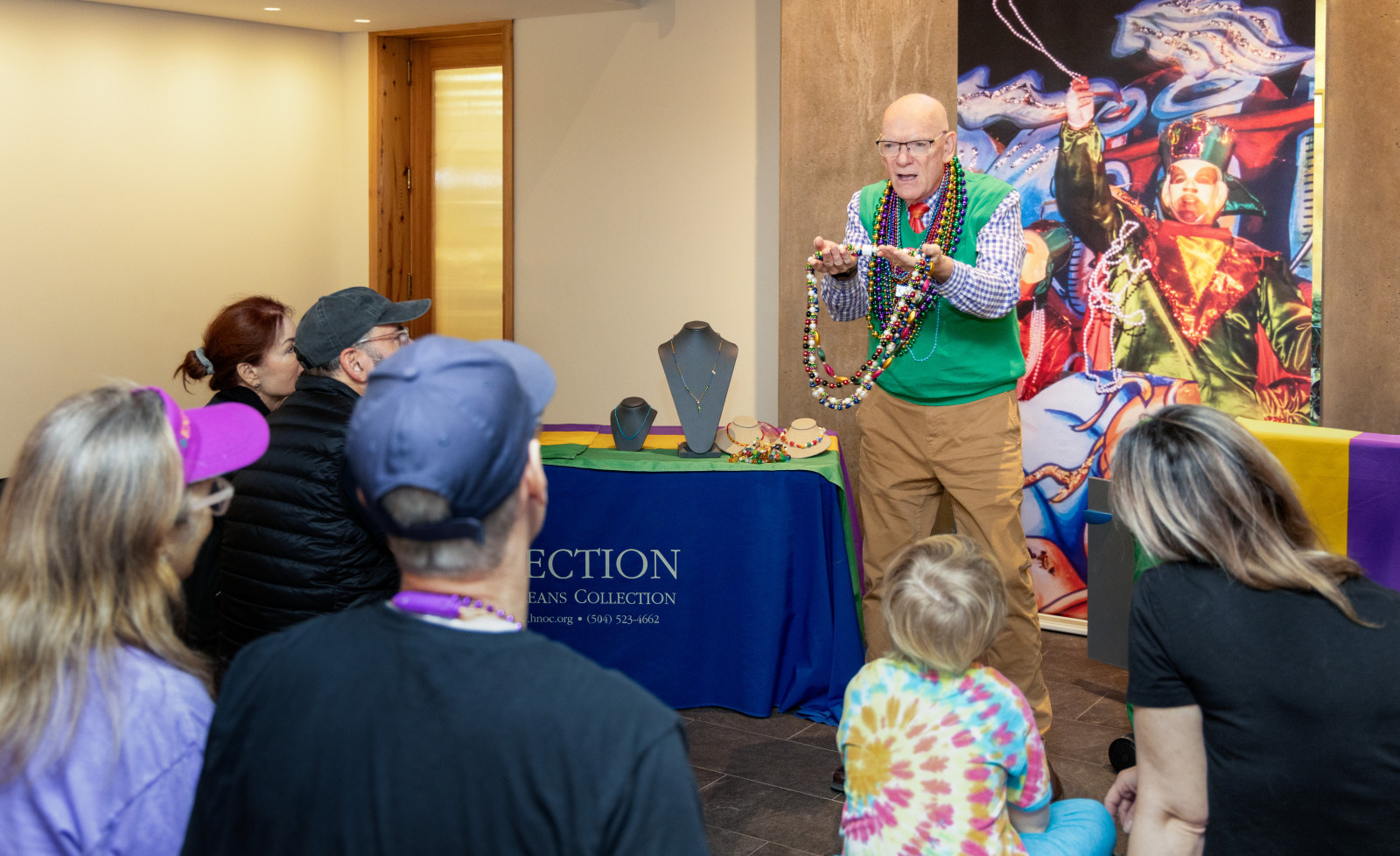 A man wearing colorful beads and gesturing animatedly stands in front of a small group seated on the floor. The room has vibrant decorations and a table with a blue cloth in the background. The group listens attentively.