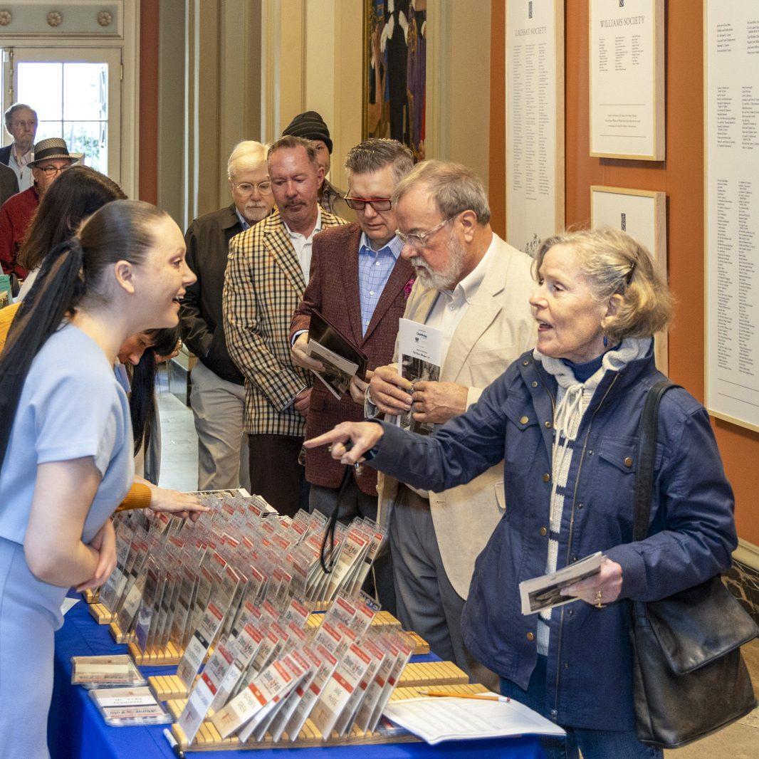People are gathered at a table covered with brochures in a hallway. A woman in a blue dress is speaking with an older woman holding a brochure. Others in line are dressed in jackets and shirts, engaging casually. Walls display framed text.