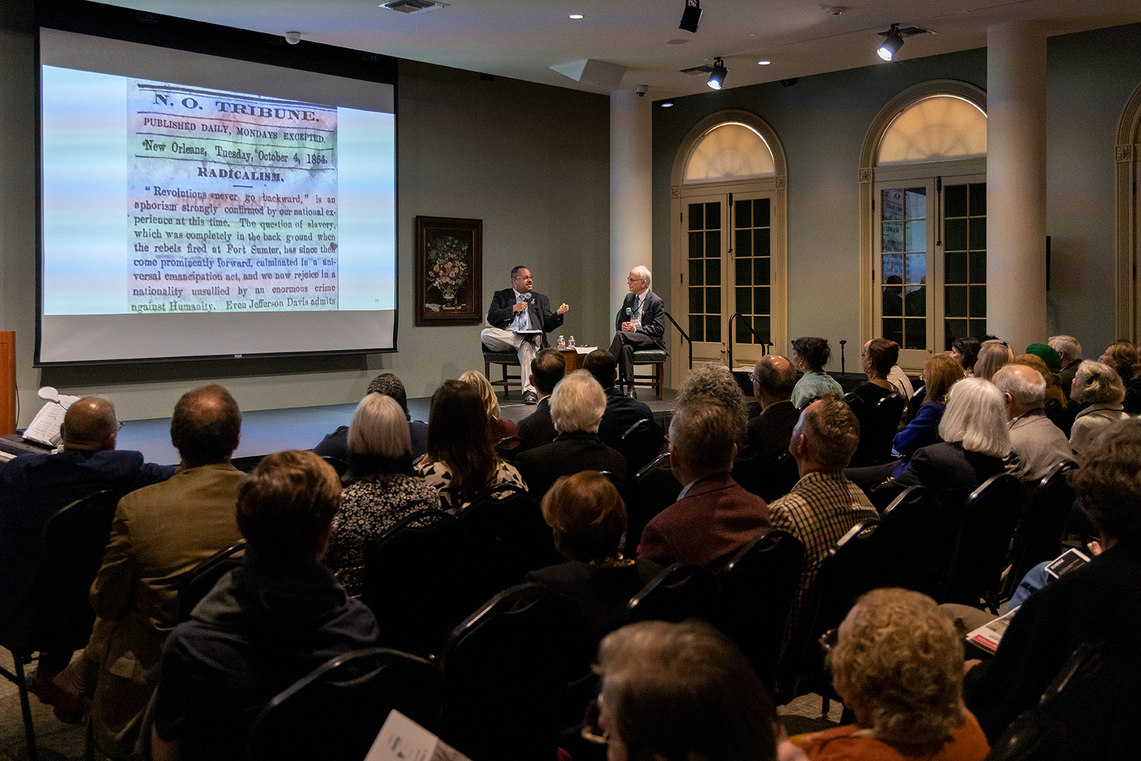 A diverse audience in a large room watches two speakers seated on stage in front of a projected historical newspaper. The room has arched windows and a screen displaying N.O. Tribune. Attendees are seated in rows, facing the speakers.
