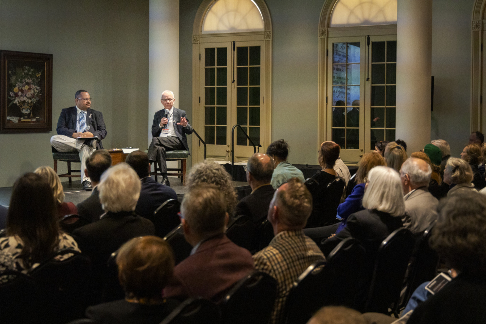 A group of people sit in an auditorium facing two speakers on stage. One speaker gestures while talking, and the other listens. Behind them, arched windows and a painting add to the settings formal atmosphere.