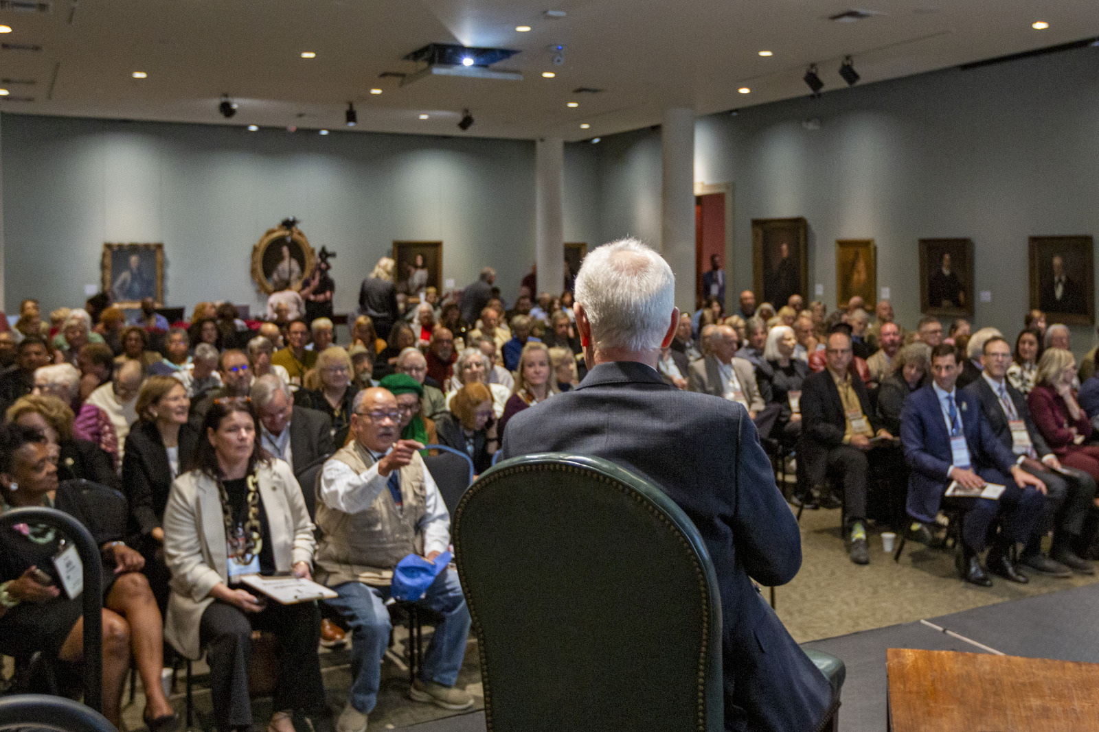 A man sits onstage facing a large audience in a gallery or conference room. The crowd is diverse, with people of various ages and backgrounds, seated and attentive. Paintings hang on the walls, adding to the formal setting.