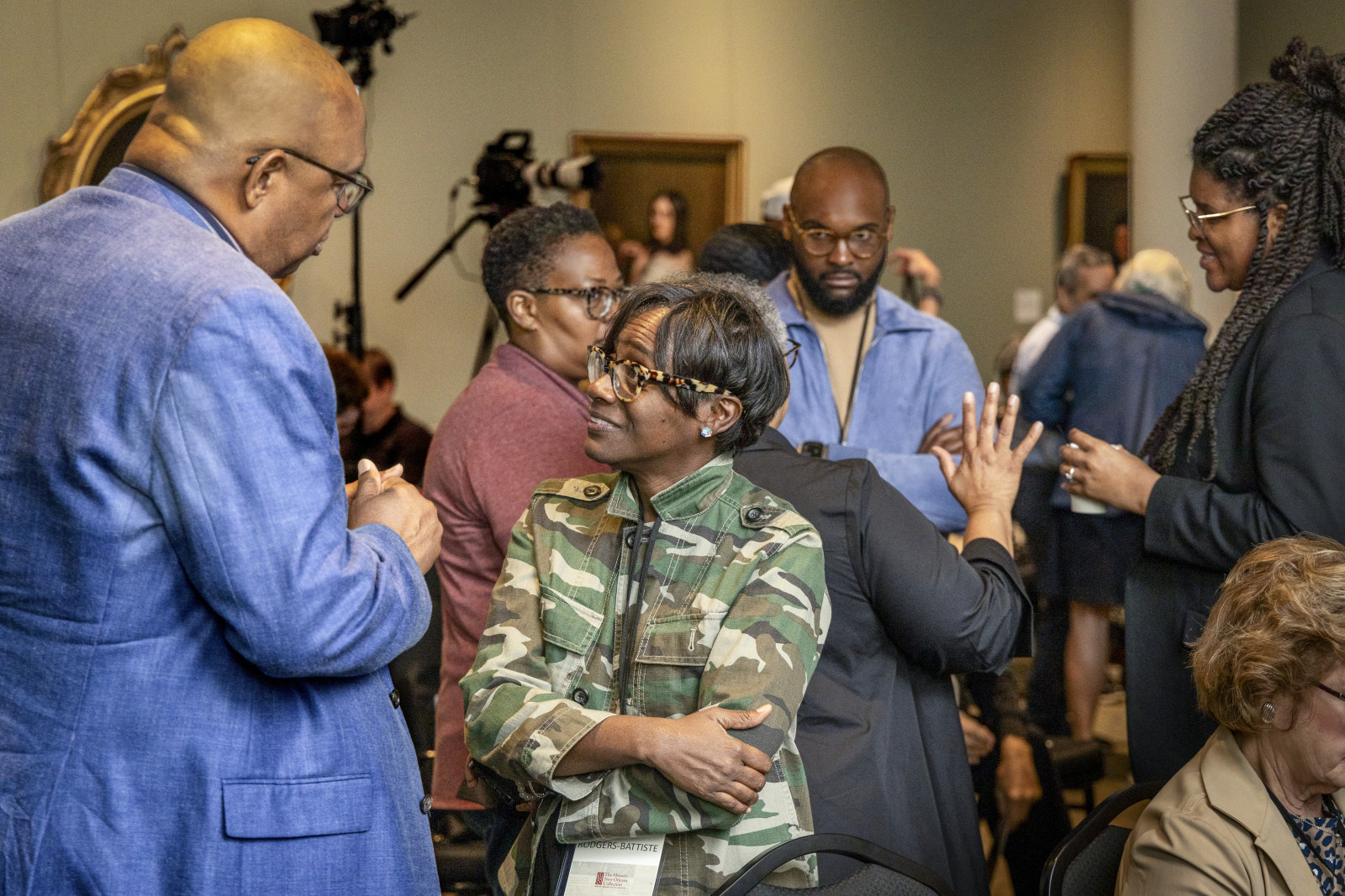 A group of people engaged in conversation at an indoor event. One person in a camouflage jacket stands with folded arms. Others are talking in pairs or groups, and there is a camera set up in the background.