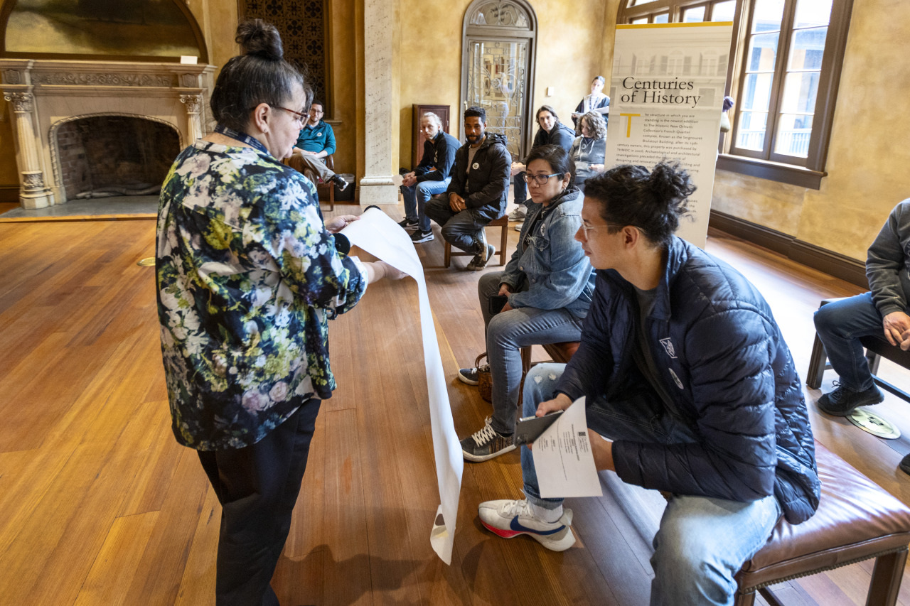 A person holds a long scroll in front of a seated group in a warmly lit room with wooden floors. A sign titled Centuries of History is in the background. The attendees are watching attentively.