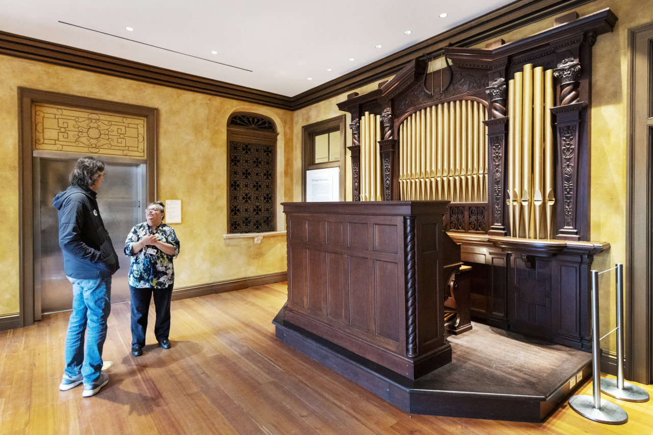 Two people stand in a room near a large, ornate pipe organ with wooden details and vertical pipes. The room has warm-colored walls and wooden flooring. One person is wearing a jacket, while the other is in a patterned shirt.