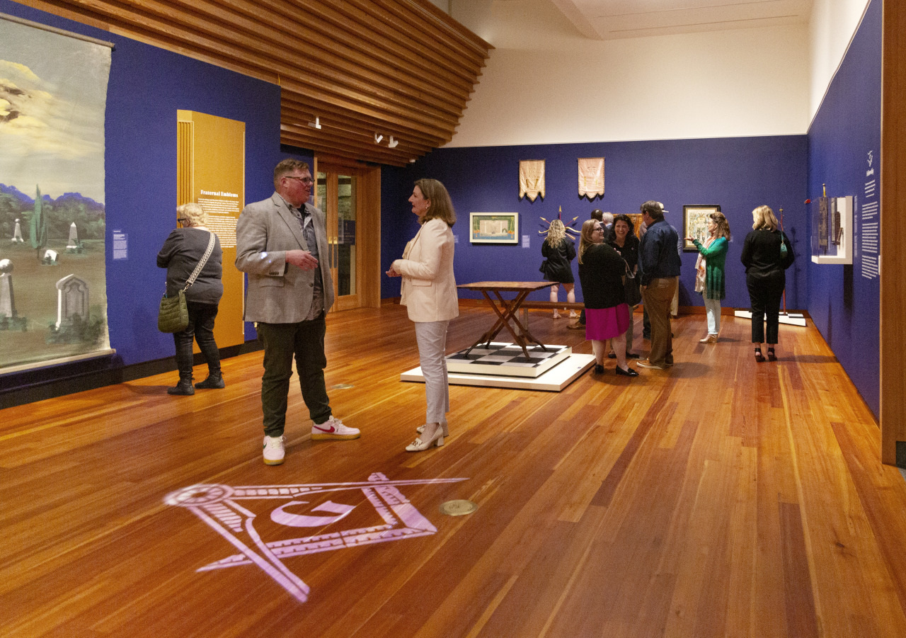 People exploring a museum gallery with wooden flooring and blue walls. Two individuals converse in the foreground. Various artworks and displays are visible on the walls and floor, including paintings and sculptures.