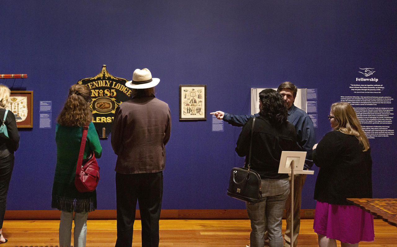 A group of people stands in an art gallery, facing a deep blue wall. They observe various framed artworks, including a large sign reading Bendix Lodge No. 85 Odd Fellows. The gallery floor is wooden, adding warmth to the scene.