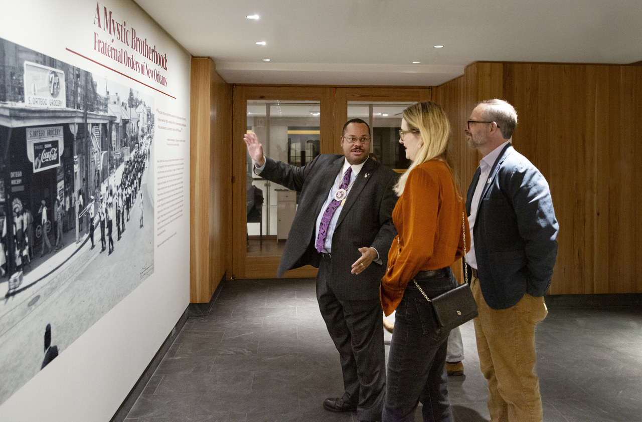 A guide gestures towards a historical photograph on the wall while speaking to a man and a woman in a gallery setting. The wall has an exhibit description titled A Most Dreadful Visitation: Frederick Douglass and The 1887 Riot.