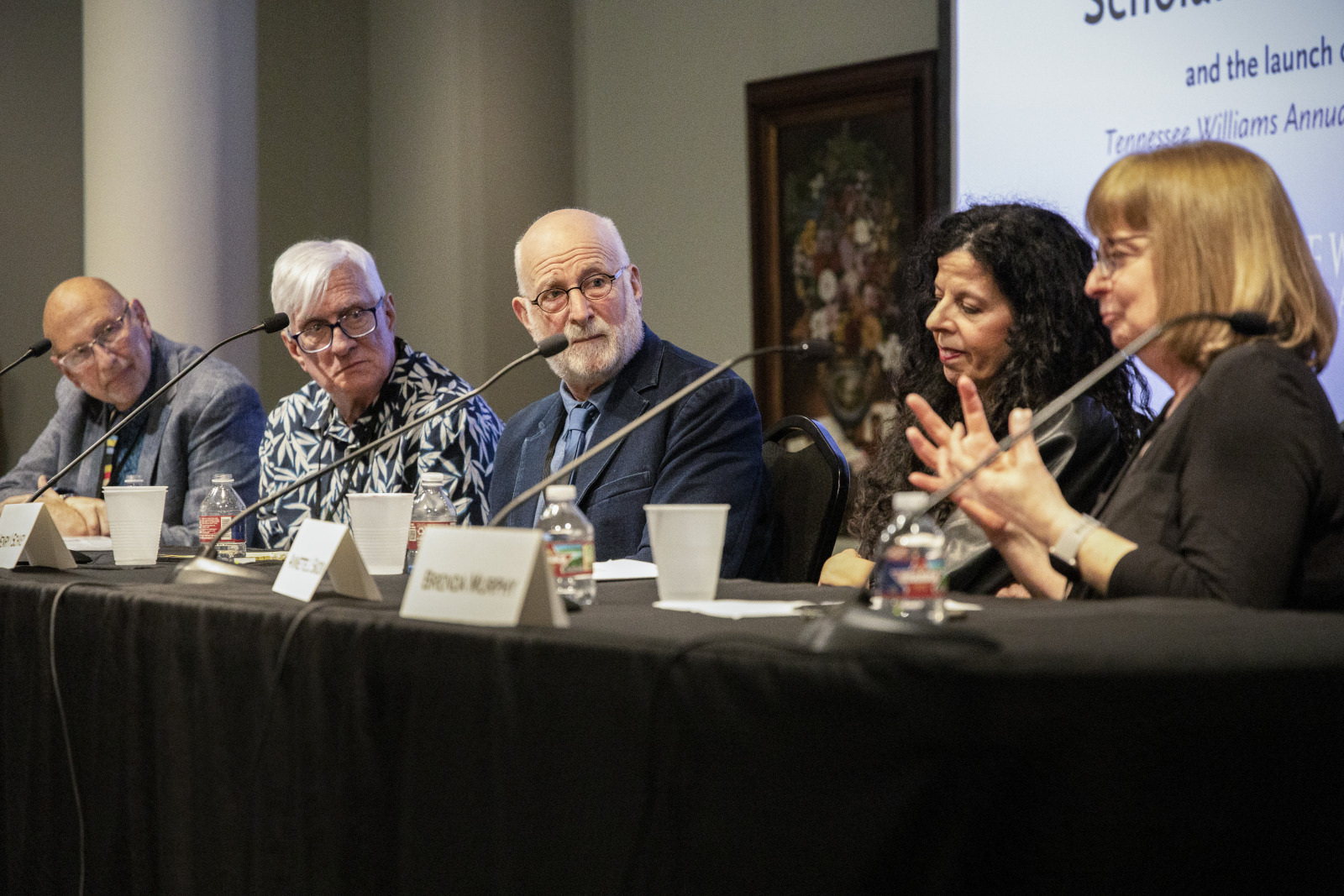 A panel of five people sitting at a table with microphones, engaging in a discussion. One woman gestures with her hands while others listen attentively. A projected screen is visible in the background.