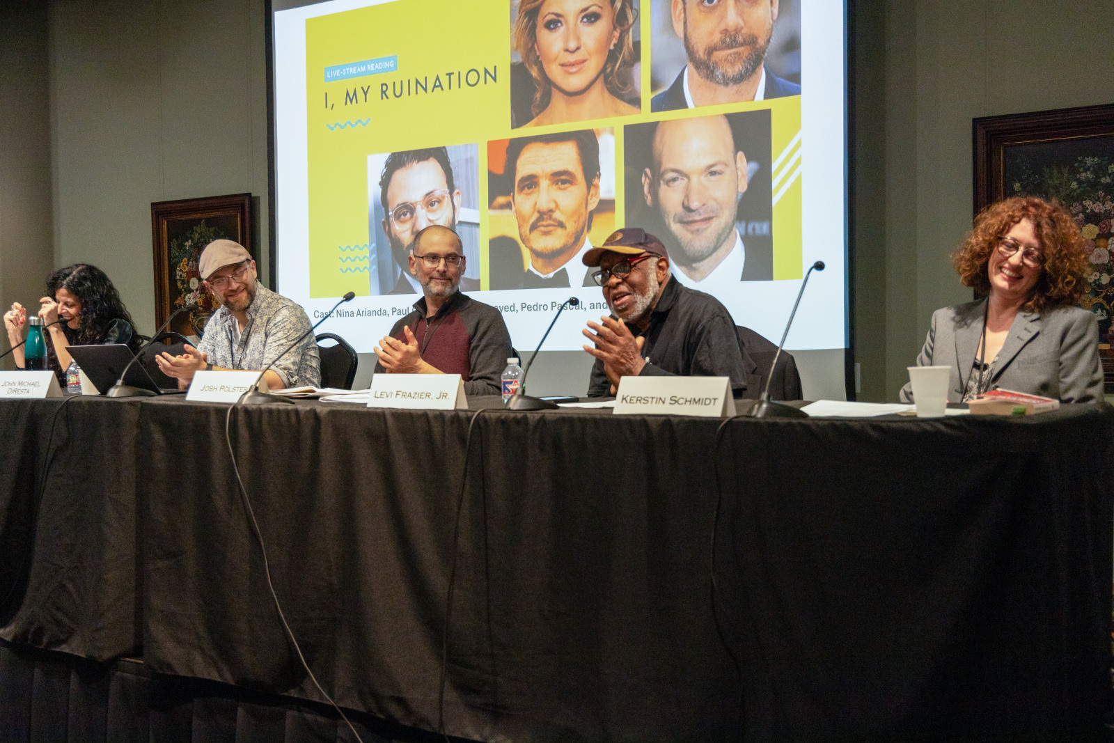 A panel discussion with six people seated at a table. They are smiling and engaged, with microphones and papers in front of them. A screen behind displays a yellow slide titled I, My Ruination featuring headshots of various people.