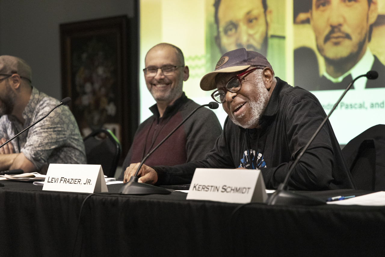 Three people sit at a panel discussion. The man closest to the camera wears a cap and glasses, smiling while speaking. The middle person is smiling and wearing glasses. Nameplates in front of them read Levi Frazier, Jr. and Kerstin Schmidt.