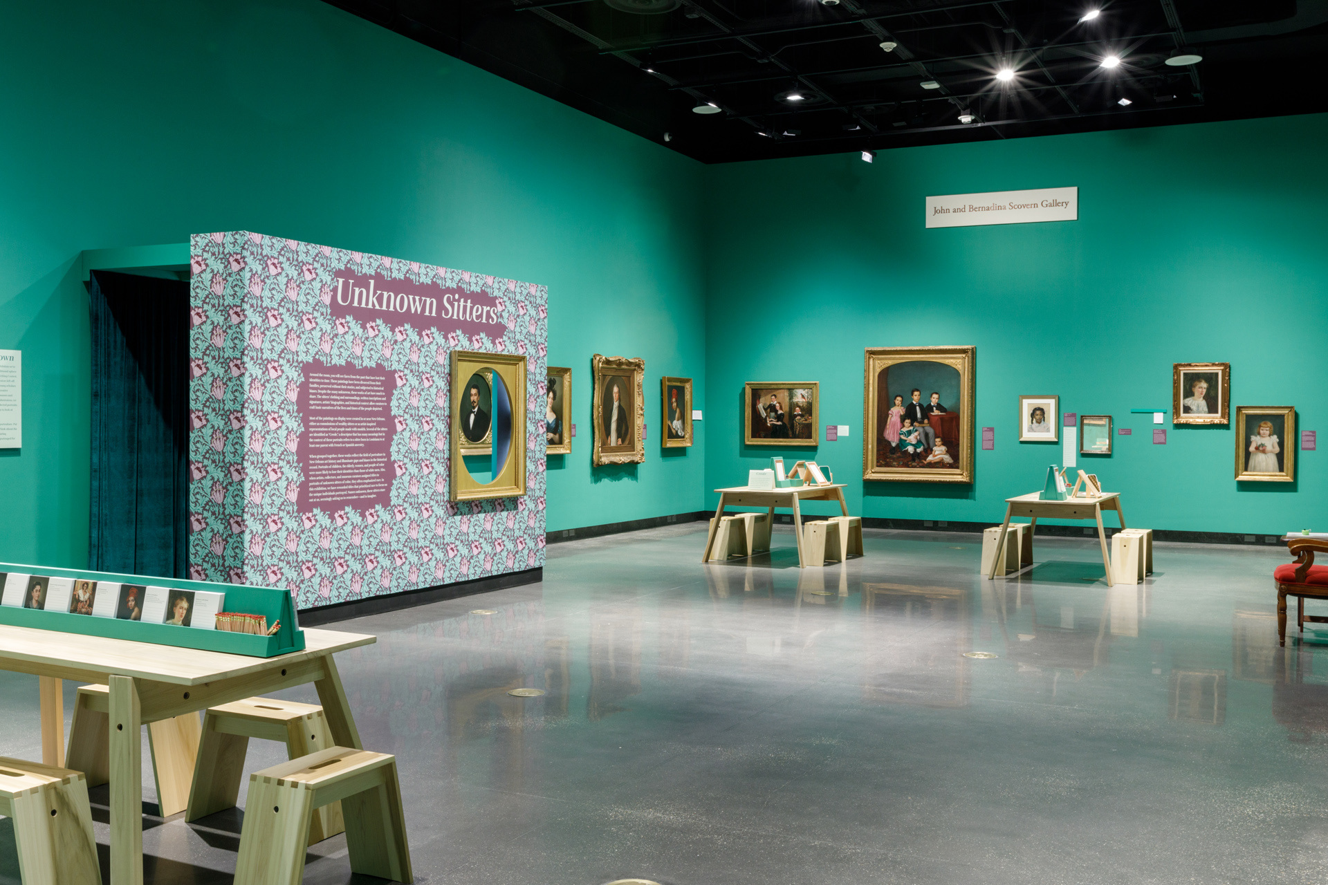 A museum gallery with teal walls displays various framed portraits. The entrance to an exhibit titled Unknown Sitters features a patterned wall. Two wooden benches and information stands are in the center of the room.