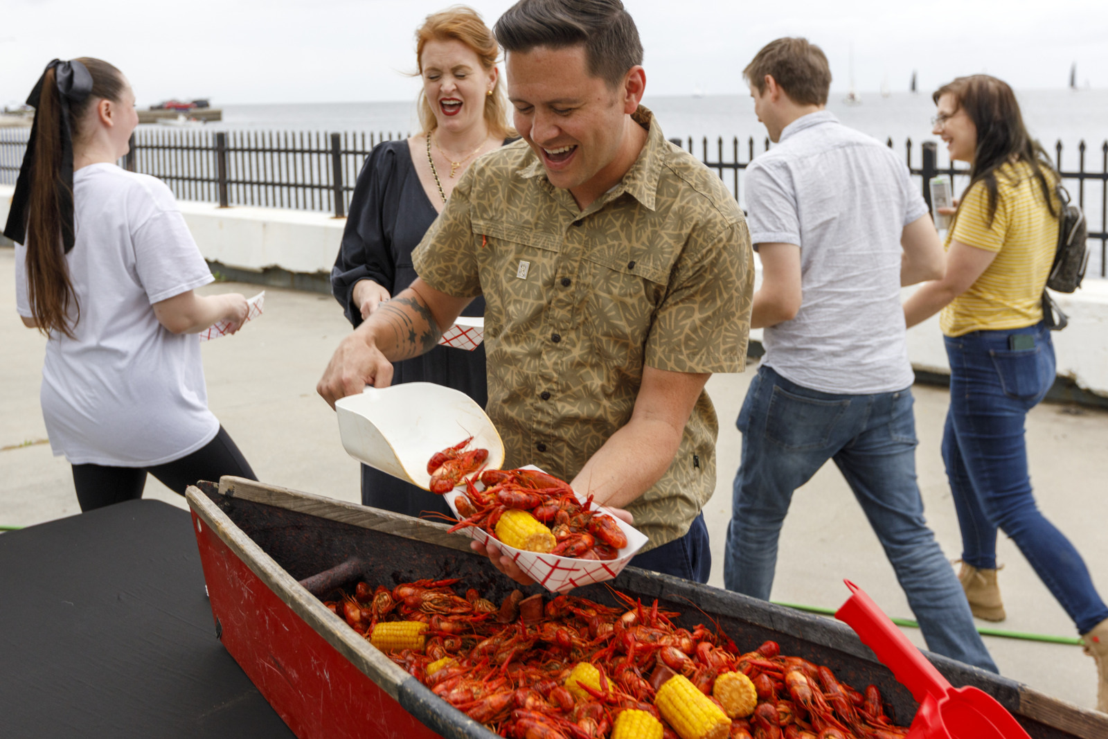 A person serves crawfish and corn from a red serving boat at an outdoor gathering by the water. Others nearby are chatting and holding food trays, enjoying the casual event.