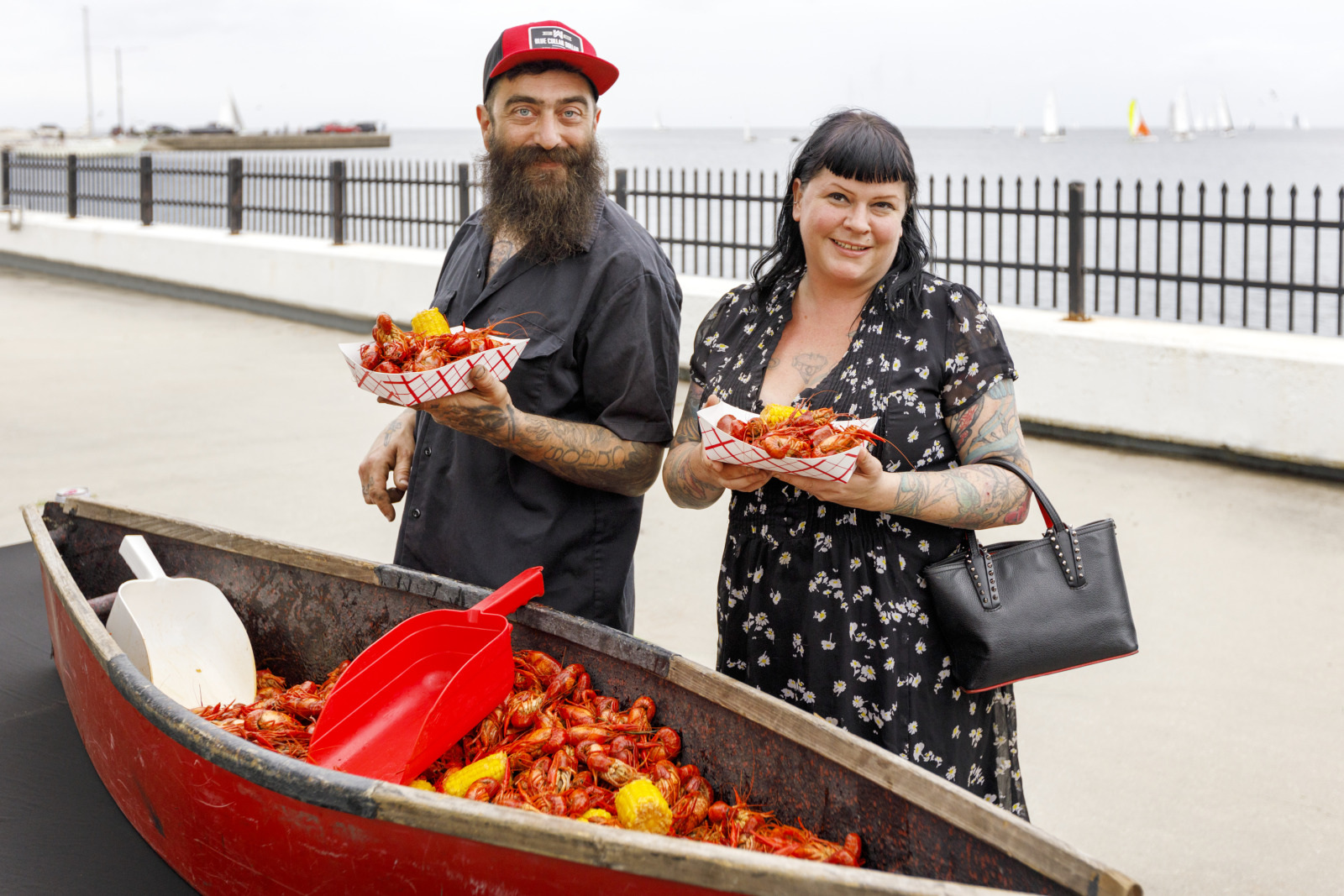 Two people smiling while holding plates of crawfish beside a red canoe filled with more crawfish. They are standing outdoors near a waterfront with sailboats in the background.