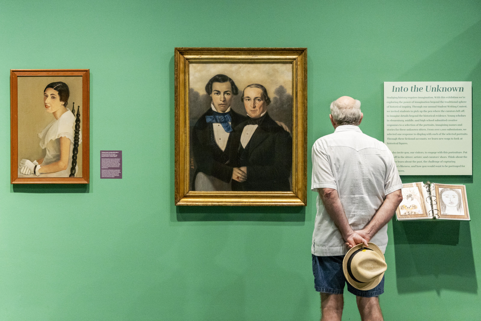 A man in casual clothing stands in front of two portrait paintings in a museum. One painting depicts a woman, and the other shows two men. The man holds a hat and reads a sign titled Into the Unknown on a green wall.
