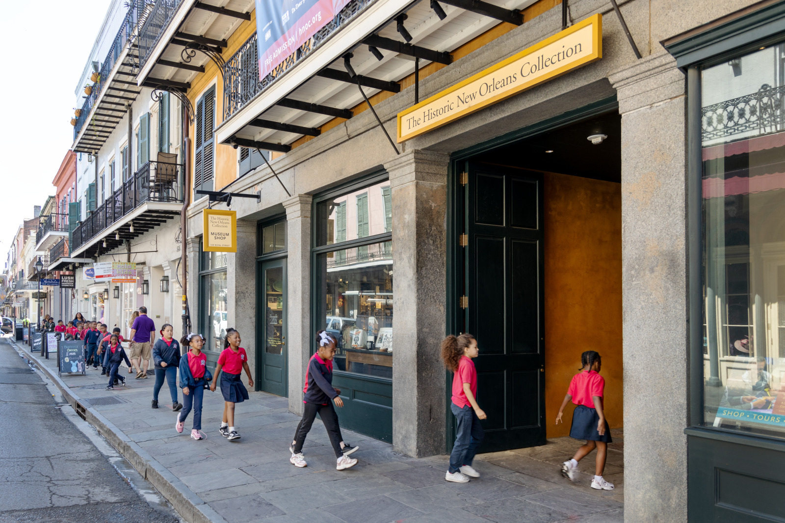 Children in red shirts walk past and enter The Historic New Orleans Collection building on a sunny street. The group is accompanied by adults. The street features historic architecture with iron balconies.