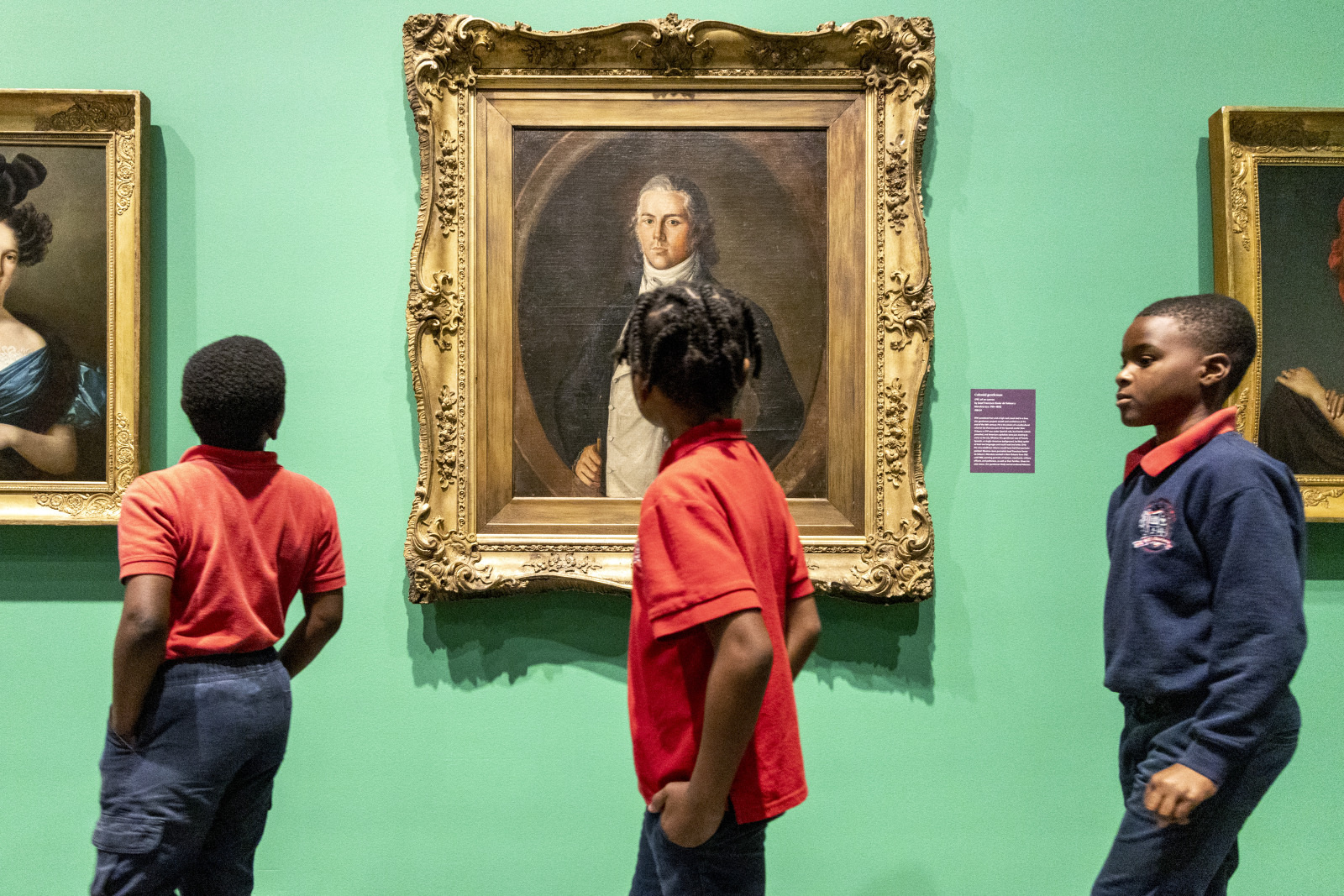 Three children in school uniforms walk past framed portraits in an art gallery with green walls. One child is looking at a specific painting, while the others walk beside him.