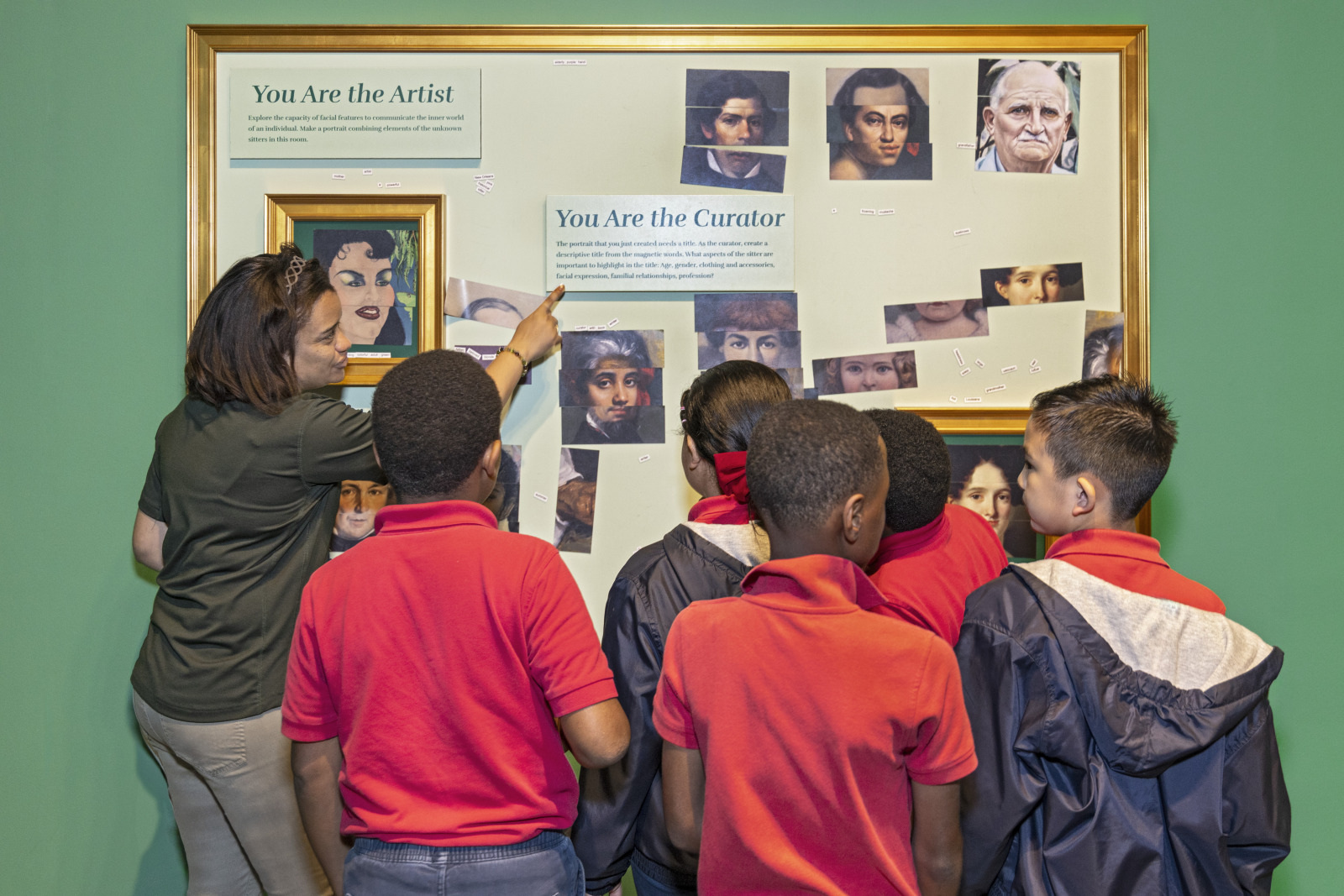 A group of children in red and dark clothing stand in front of an art exhibit. A woman points at the display, which features various portraits and text labeled You Are the Artist and You Are the Curator. The background is green.