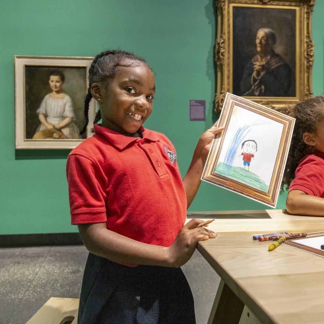 A young girl in a red polo shirt smiles proudly while holding up her drawing in a museum. Behind her, classic paintings adorn the green wall. Crayons and paper are on the table in front of her.
