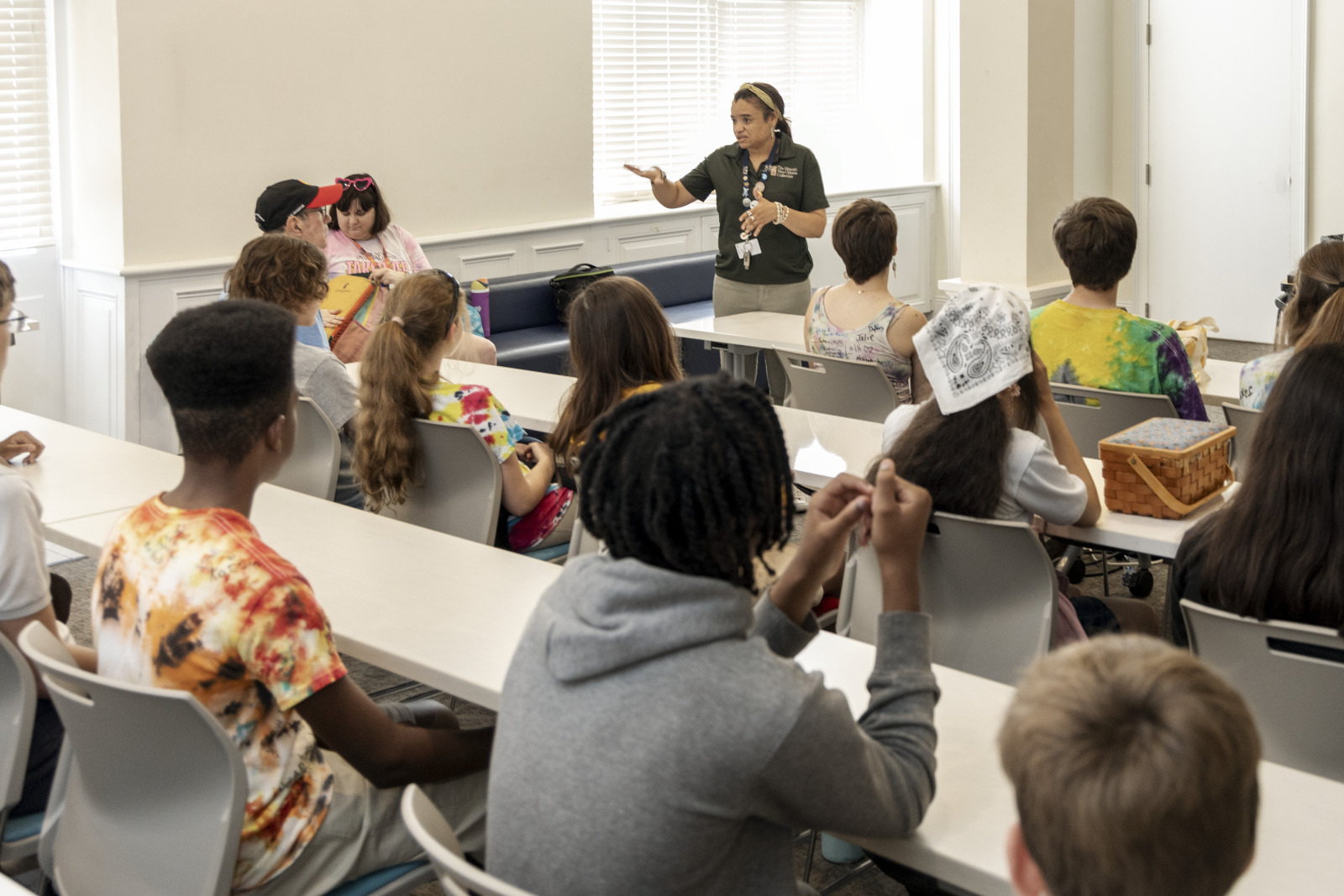 A group of students sit in a classroom with a teacher standing at the front. The students are listening attentively. The room is bright with natural light coming through the windows.