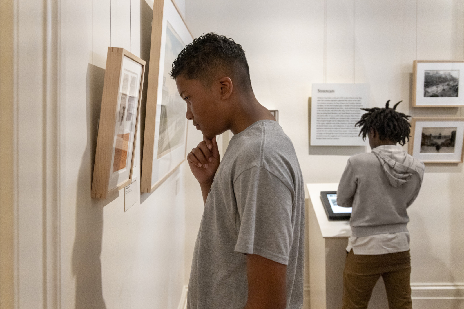 Two people in a gallery: one person observes framed art, another interacts with a digital display. The walls showcase various artworks and descriptions.