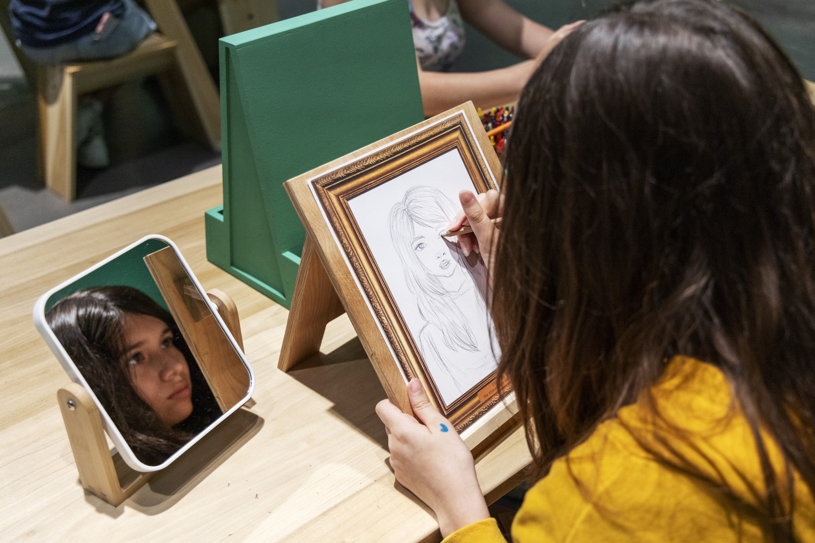 A person with long hair sketches a portrait at a wooden desk. A small mirror rests on the table, reflecting their face. The person is wearing a yellow sweater, and art supplies are scattered on the table.