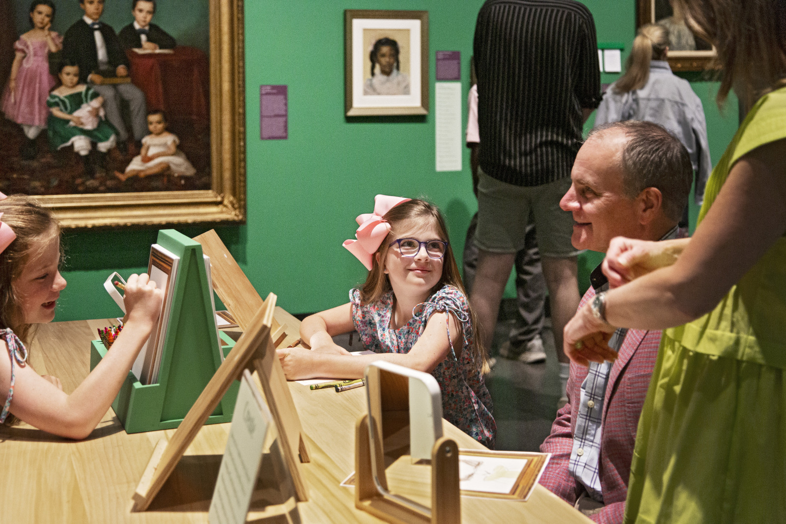 A family enjoys an art museum, sitting at a table with drawing boards. Two children, wearing bows and glasses, draw while two adults accompany them. Framed paintings are on the green walls in the background.